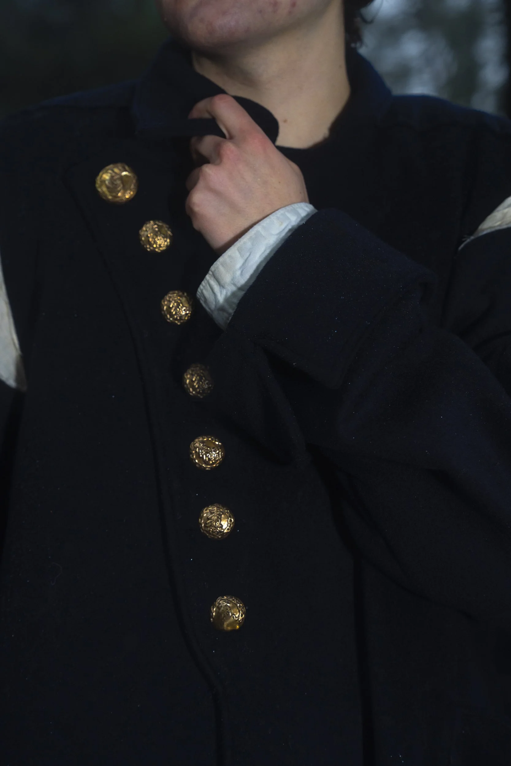 Close-up of a person's neck and hand adjusting the collar of a formal black coat with gold buttons and decorative pins. The coat is worn over a white shirt.