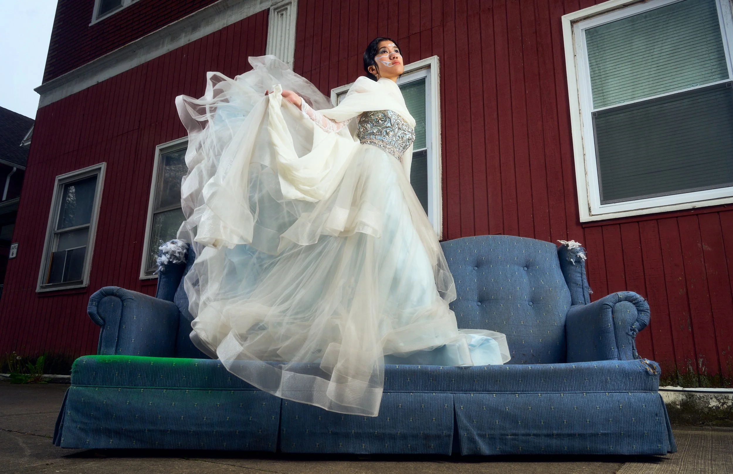 Woman dressed in a white ornate gown standing on a blue couch outside in front of a red wooden building with large windows.