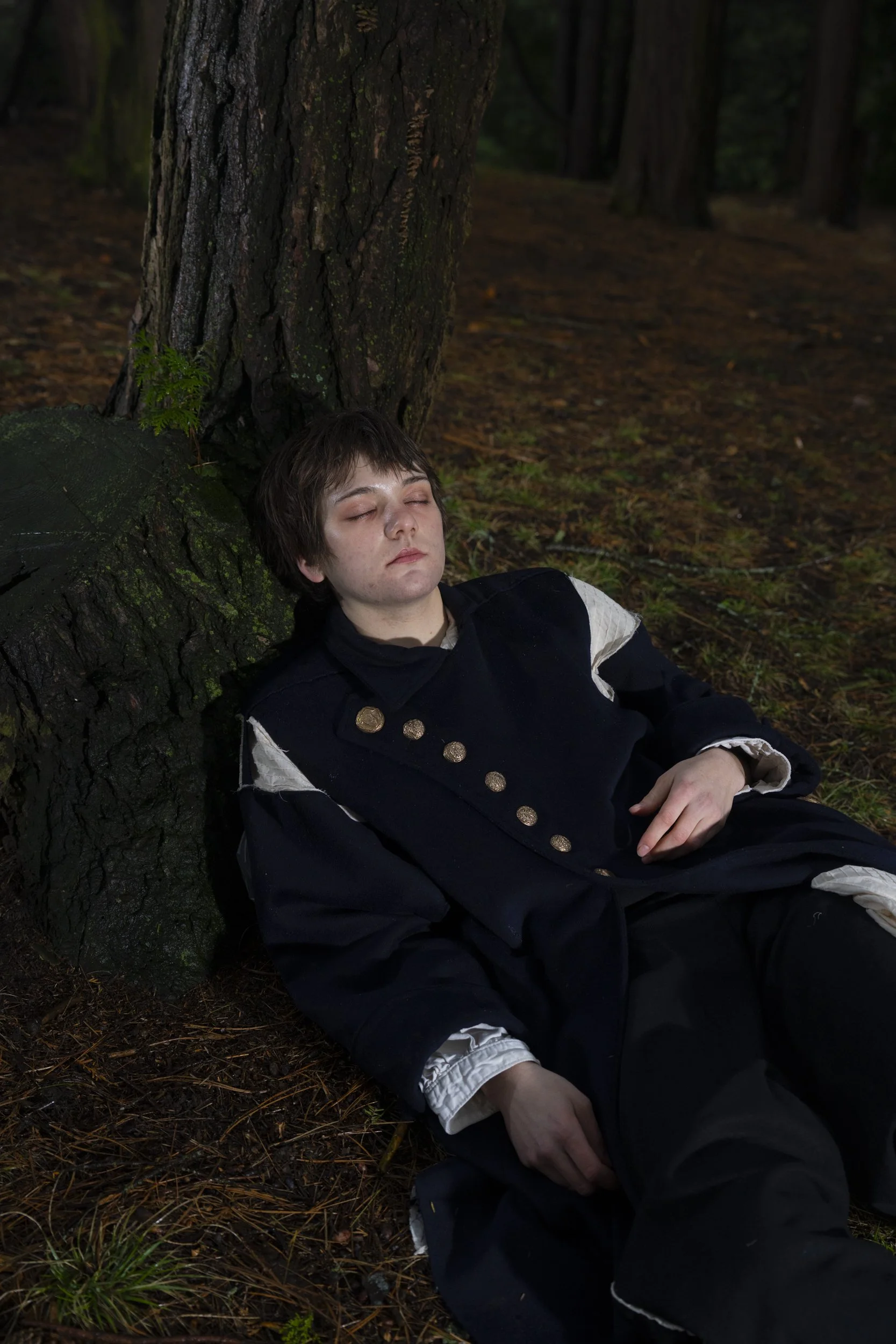 A young person with short brown hair sleeps against a large tree in a forest. They are wearing historical clothing with decorative buttons and light-colored undershirts. The forest floor is covered with pine needles and small plants, and tall trees a