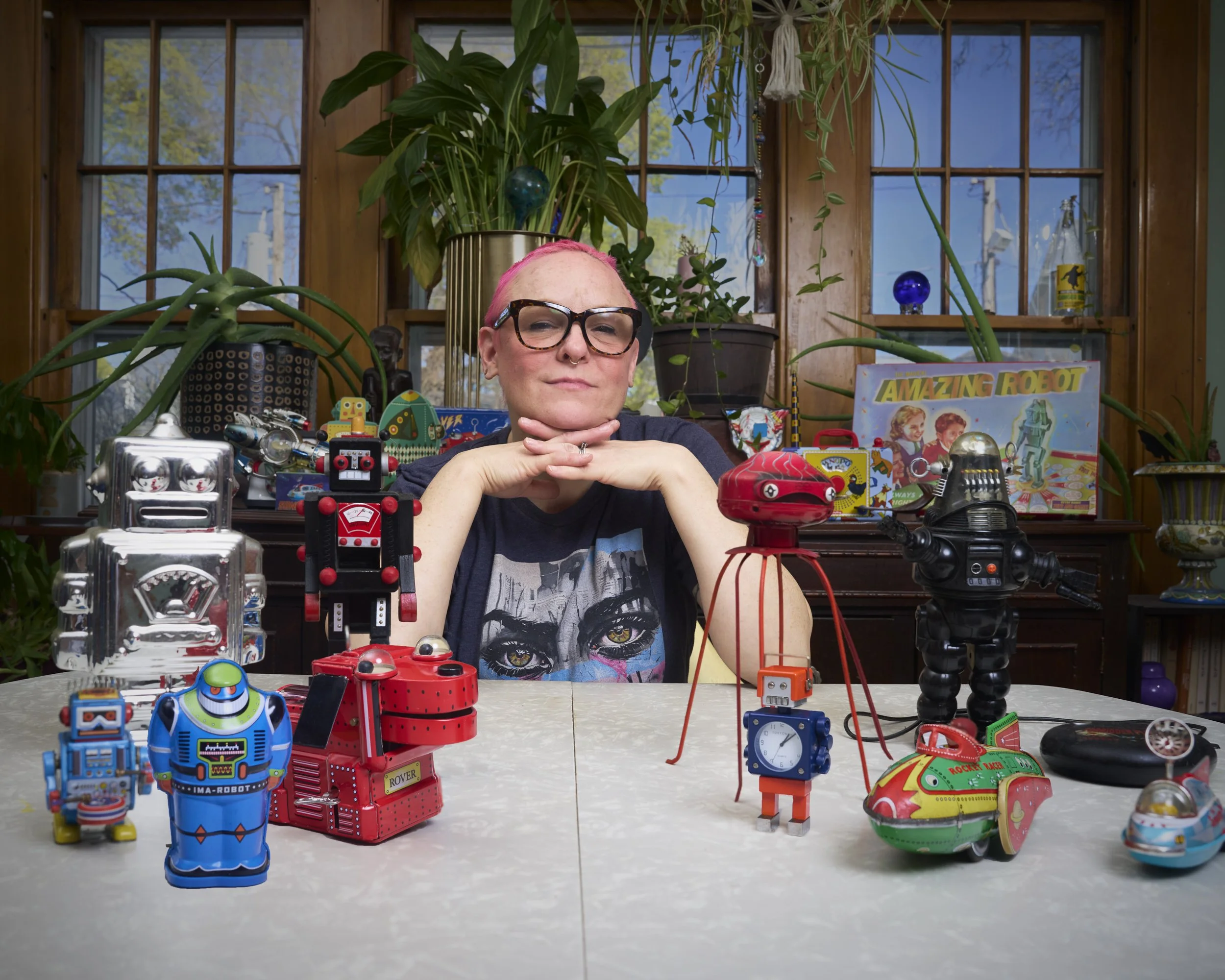 A person with pink hair and glasses sits at a table with various vintage robot toys and collectibles displayed in front of them, inside a room with large windows and lots of houseplants.
