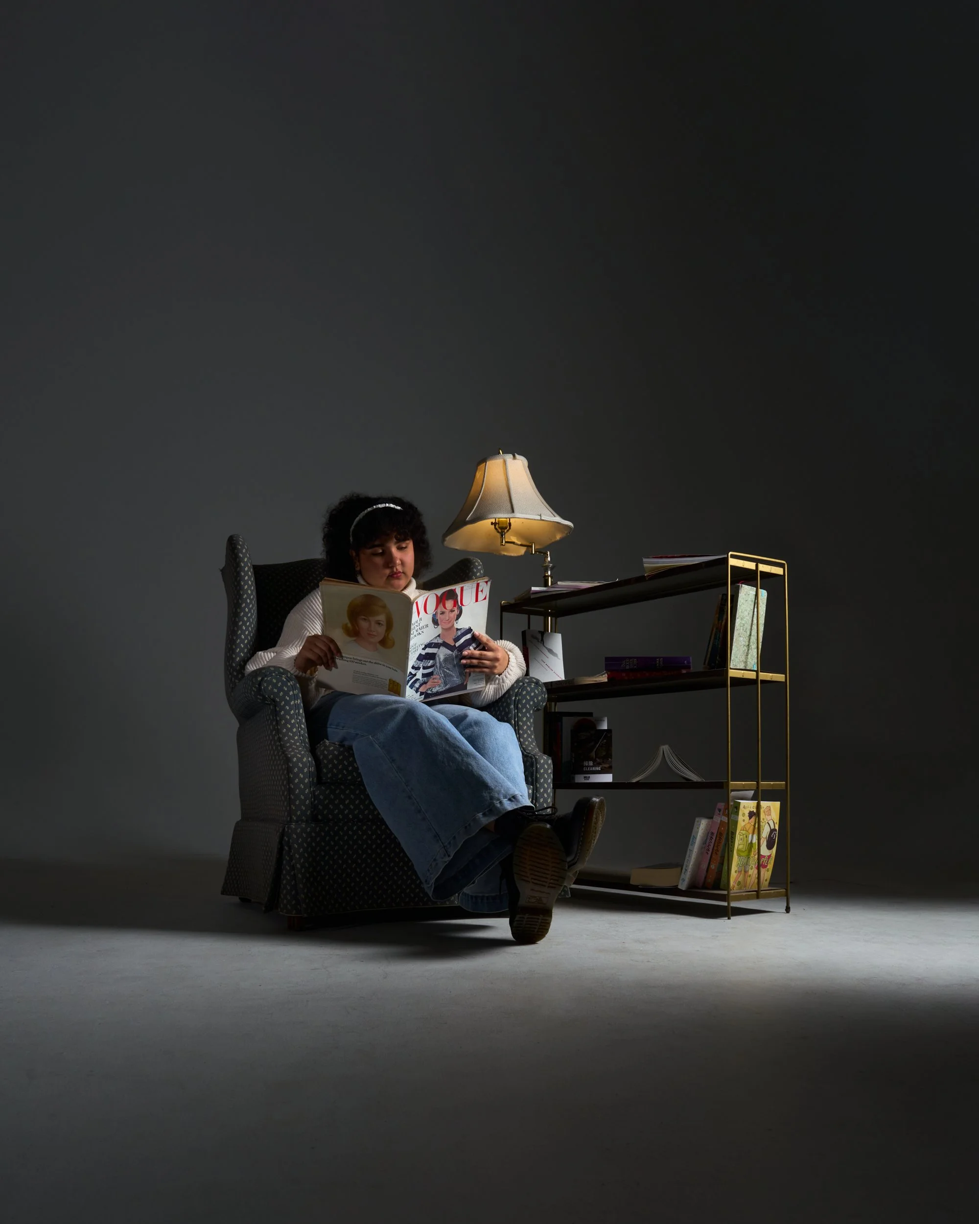 A young girl is sitting in an armchair, reading a copy of Vogue magazine. A small bookshelf with a gold frame holding more books and magazines is nearby. A lamp with a beige shade provides light. The background is a plain dark wall.