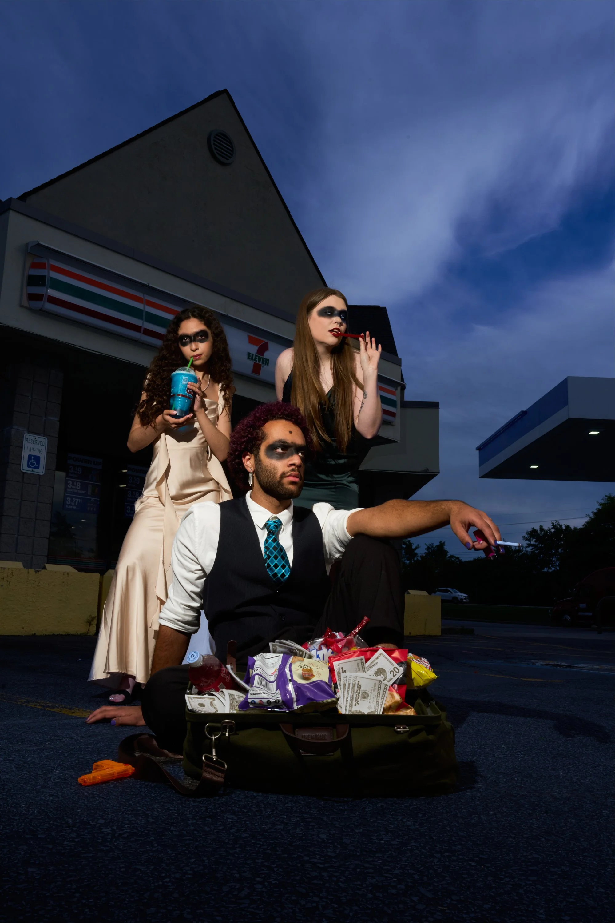 young people dressed in formal attire, with dark makeup and white face paint resembling masks, are posing outside a 7-Eleven store during twilight. One person is holding a soda, another is biting into a red licorice stick, and the third is sitting on