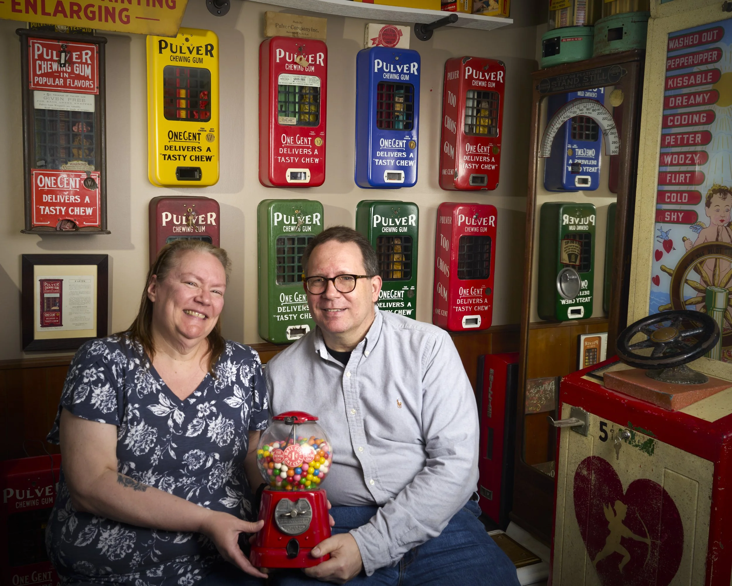 A smiling woman with glasses and a smiling man sitting next to each other, holding a red gumball machine filled with colorful gumballs in a room with vintage candy and gum vending machines on the wall behind them.
