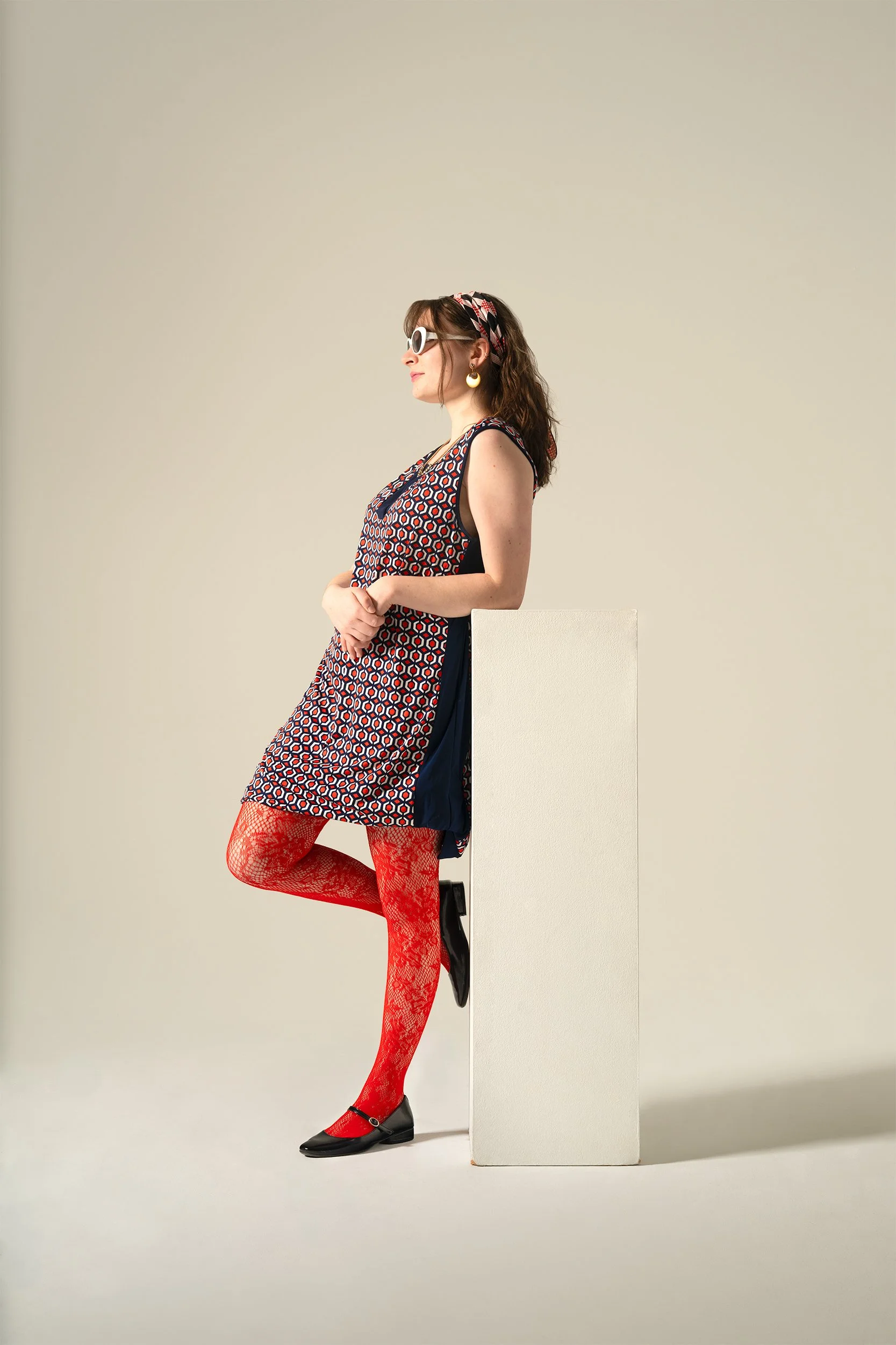 Side view of a woman wearing large white sunglasses, a patterned dress, red lace tights, black shoes, with a headband, earrings, and posing against a plain background, with one foot resting on a white block.