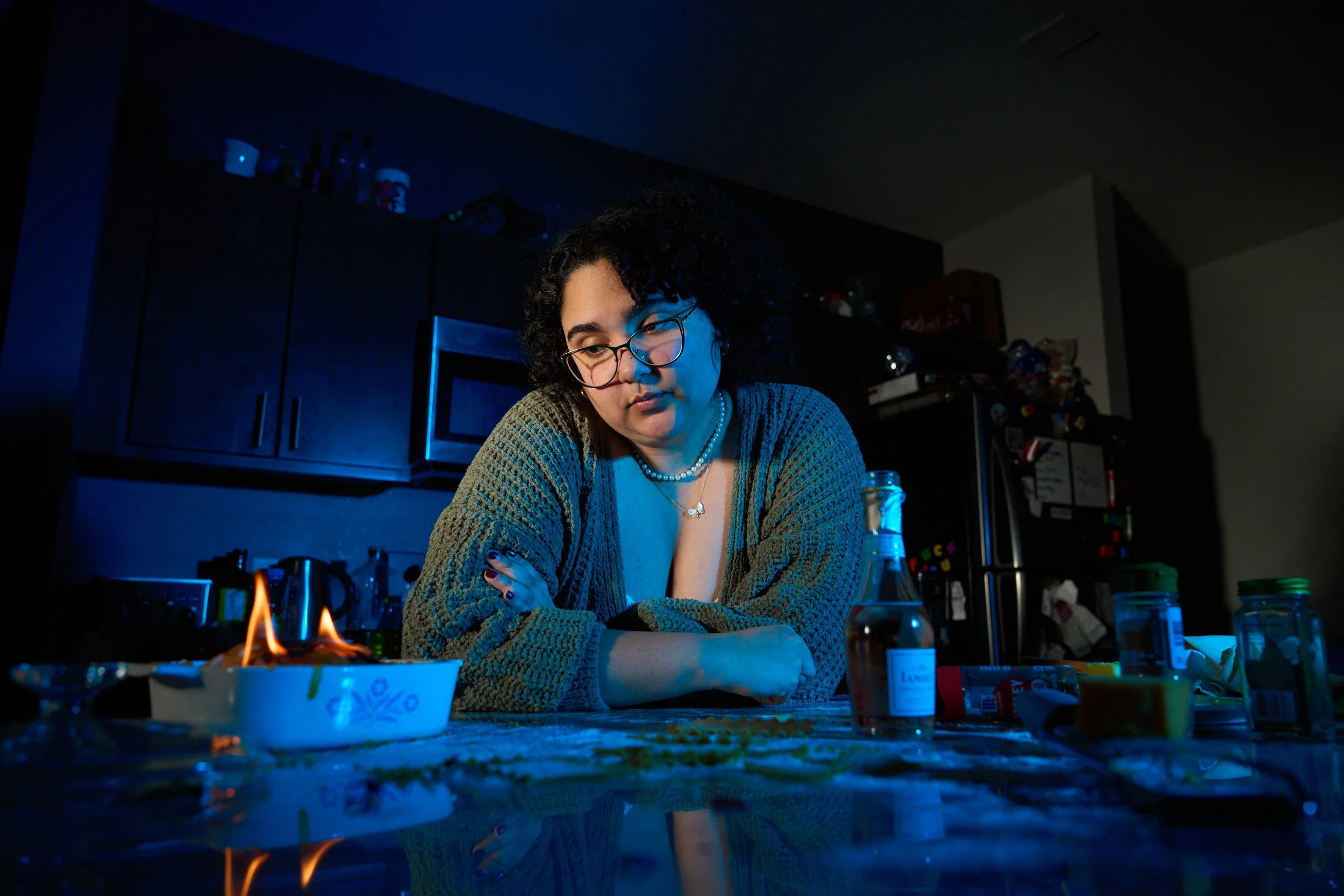 A woman with curly dark hair, glasses, and a pearl necklace is sitting at a kitchen counter with a blue lighting. She appears to be in thought, surrounded by various bottles of spices and liquor, with a small casserole dish on fire in front of her.