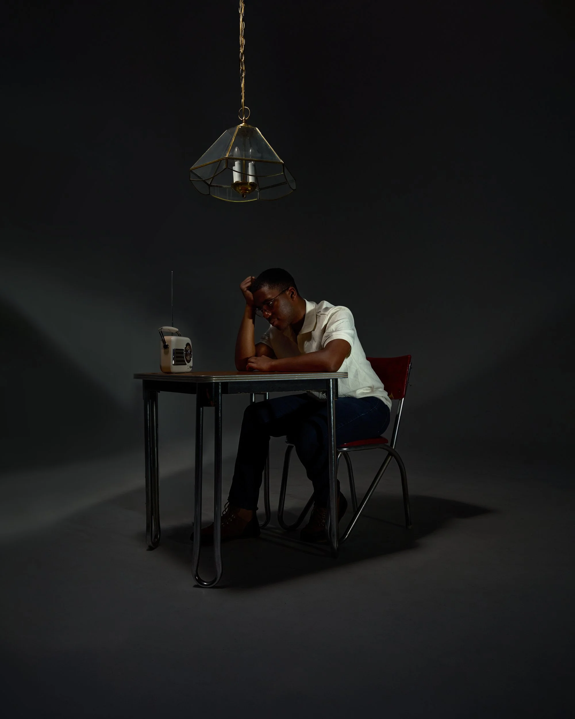 A young man wearing glasses sitting at a table, resting his head on his left hand, with a vintage radio on the table and a pendant light hanging above him against a dark background.