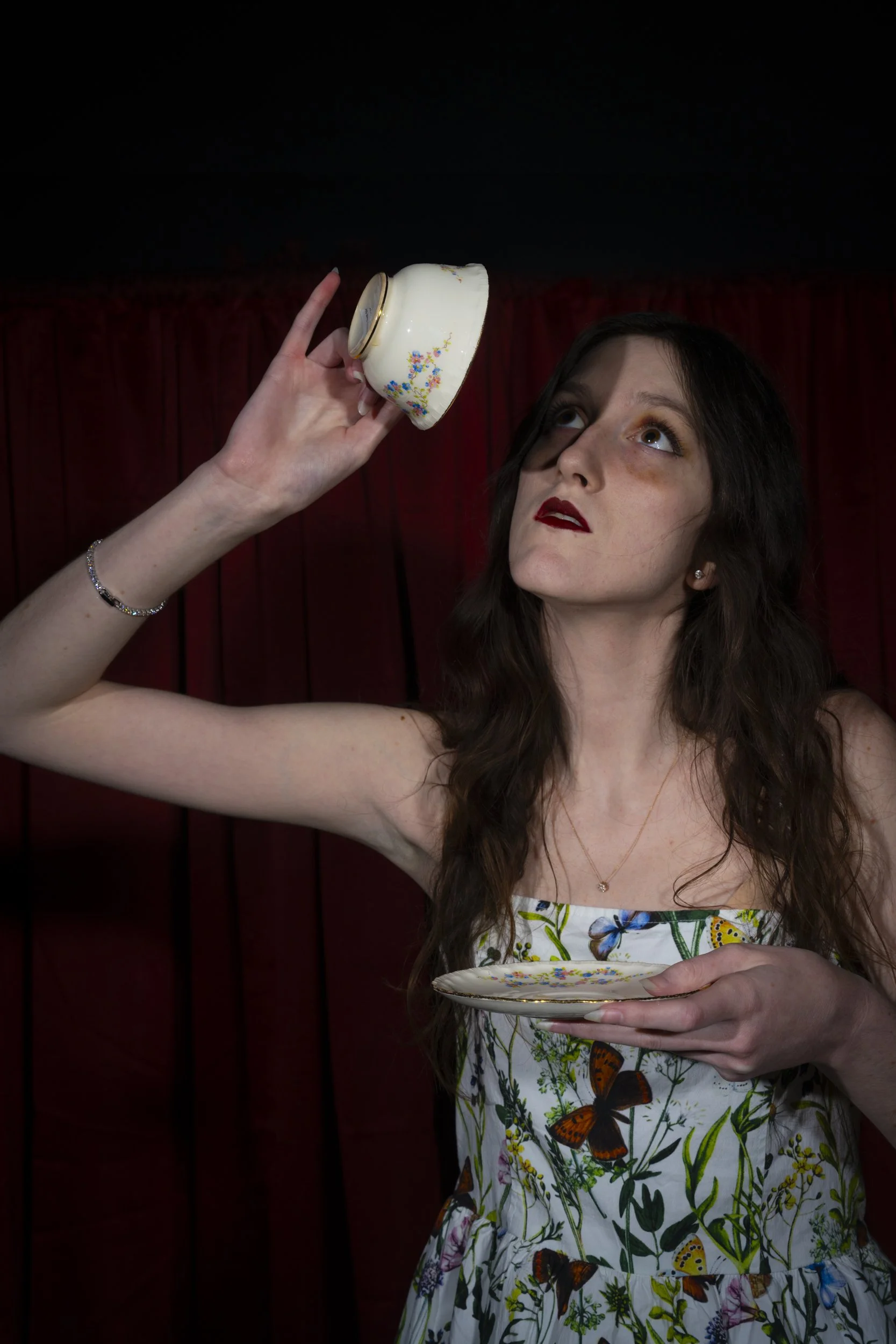 A young woman with long brown hair and dark lipstick holds a small, white teacup decorated with colorful flowers and a matching saucer. She is wearing a strapless dress with a butterfly and floral pattern. Her gaze is upwards, and she is standing in 