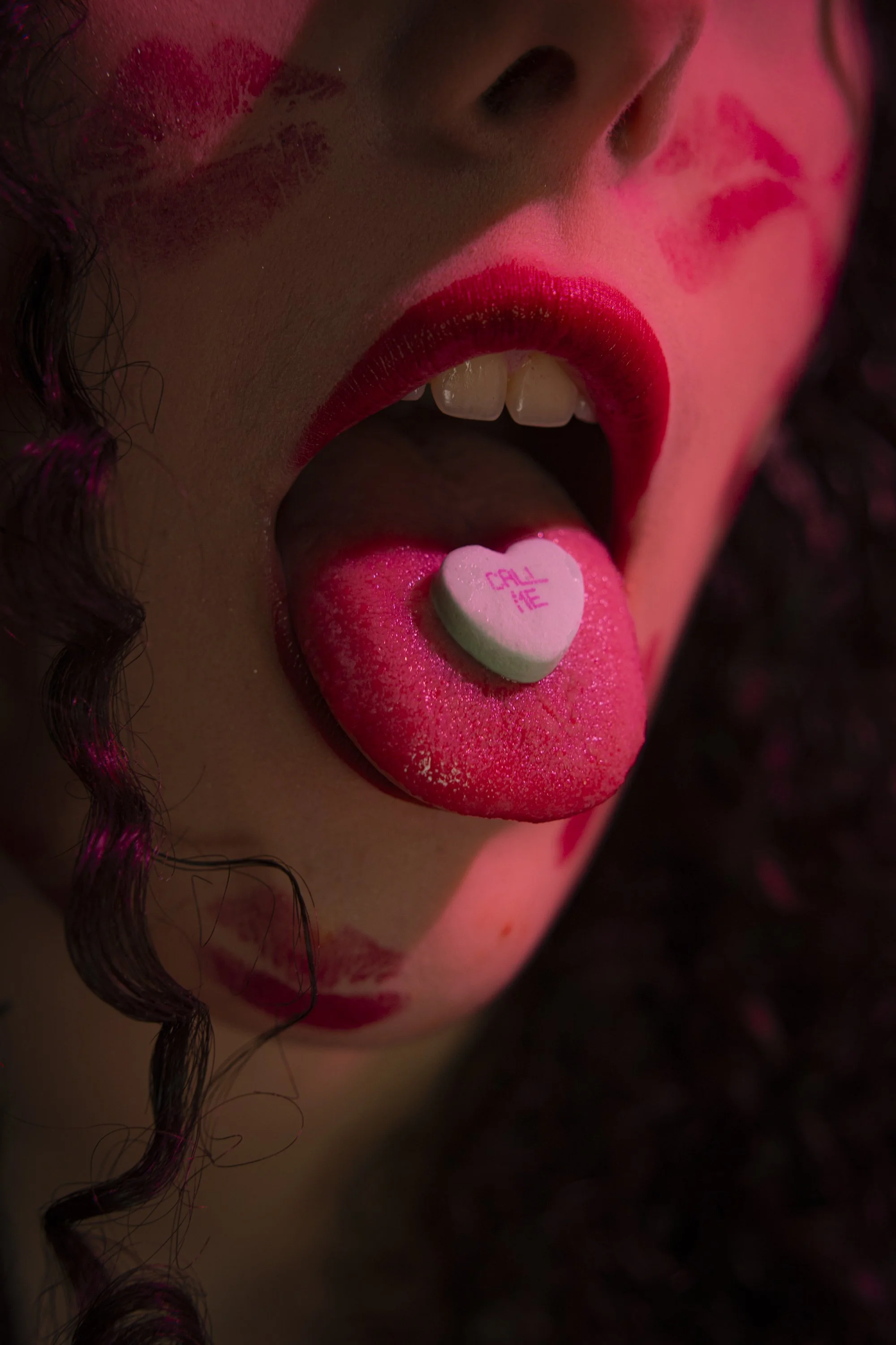 Close-up of a woman's mouth with bright red lipstick, her tongue extended holding a white candy heart with pink writing that says 'CALL ME'.