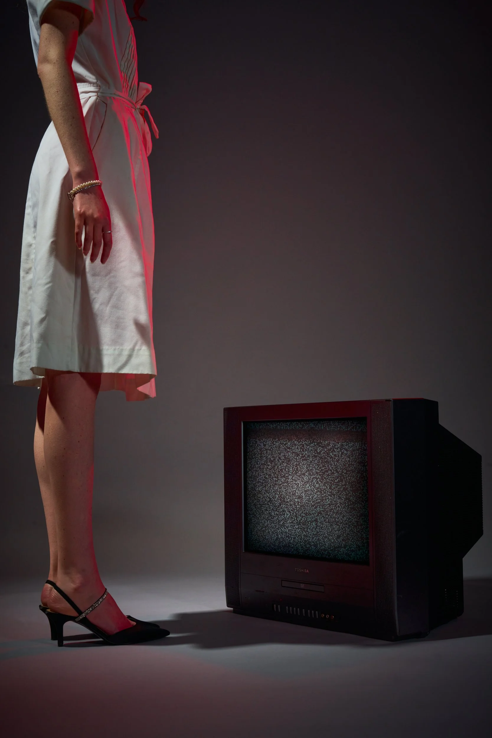 A woman in a white dress and high heels stands next to an old television with static on the screen, against a dark background.