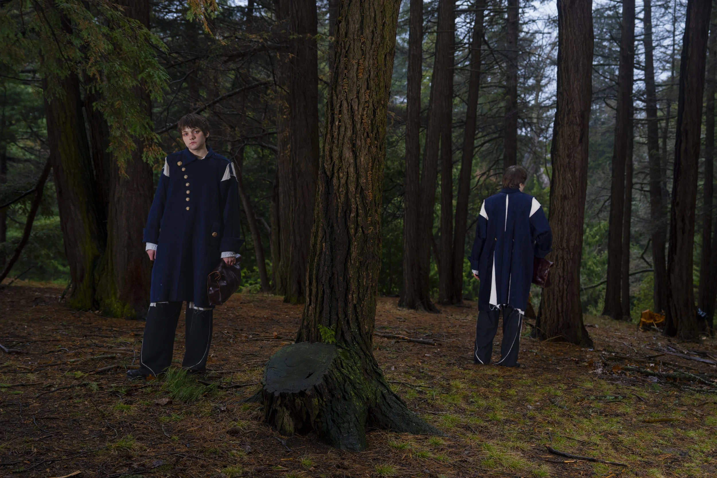 Two boys dressed in dark blue coats standing in a dense forest of tall trees with green foliage. One boy faces forward while the other faces away.