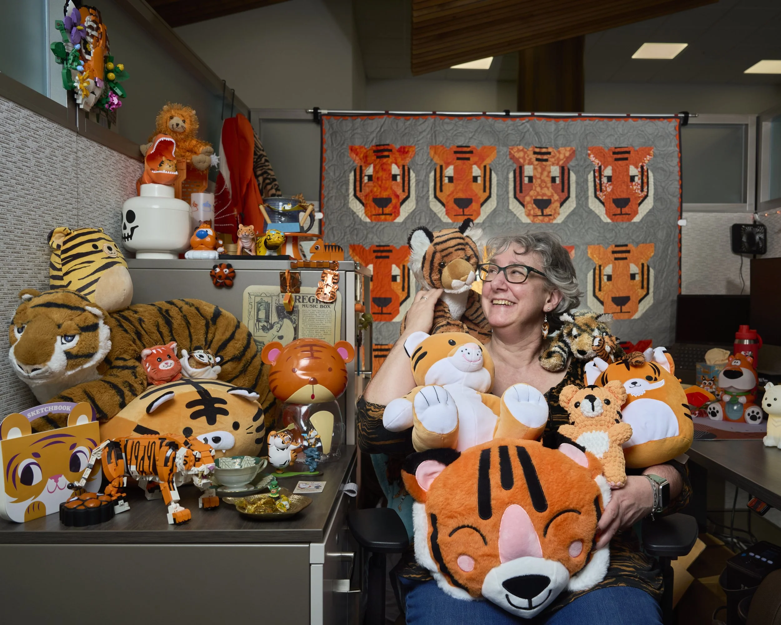 A woman with glasses smiling and holding multiple plush tiger toys, surrounded by various tiger-themed stuffed animals and toys on a desk and shelf, with a tiger face quilt hanging behind her and tiger plush toys displayed around her.