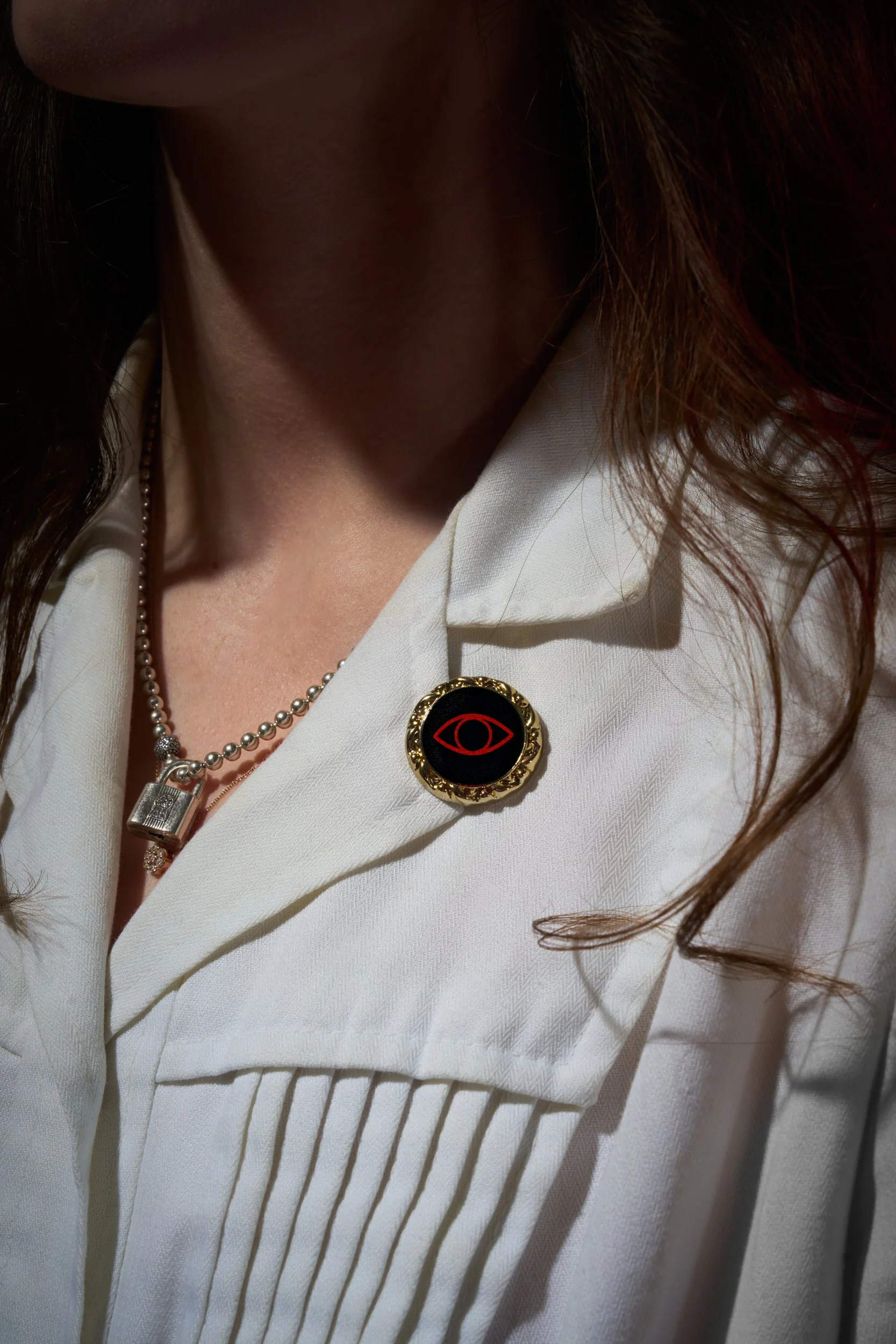 Close-up of a woman wearing a white blouse with a decorative black and gold brooch and a pearl necklace, with long brown hair partially covering her face.