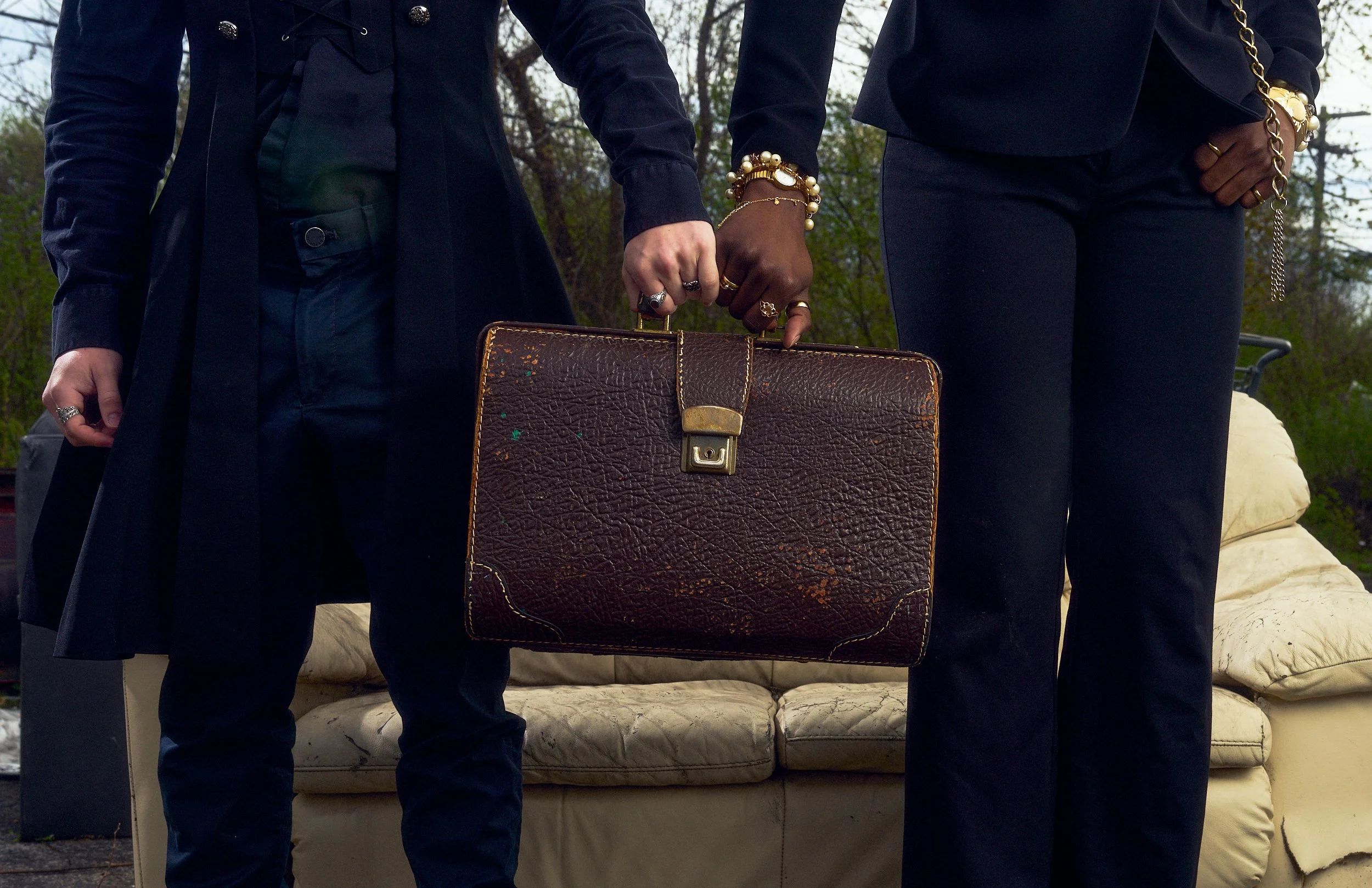 Two people standing outdoors, dressed in dark clothing, holding a vintage brown leather suitcase between them. The focus is on their hands and the suitcase, with trees and a couch in the background.