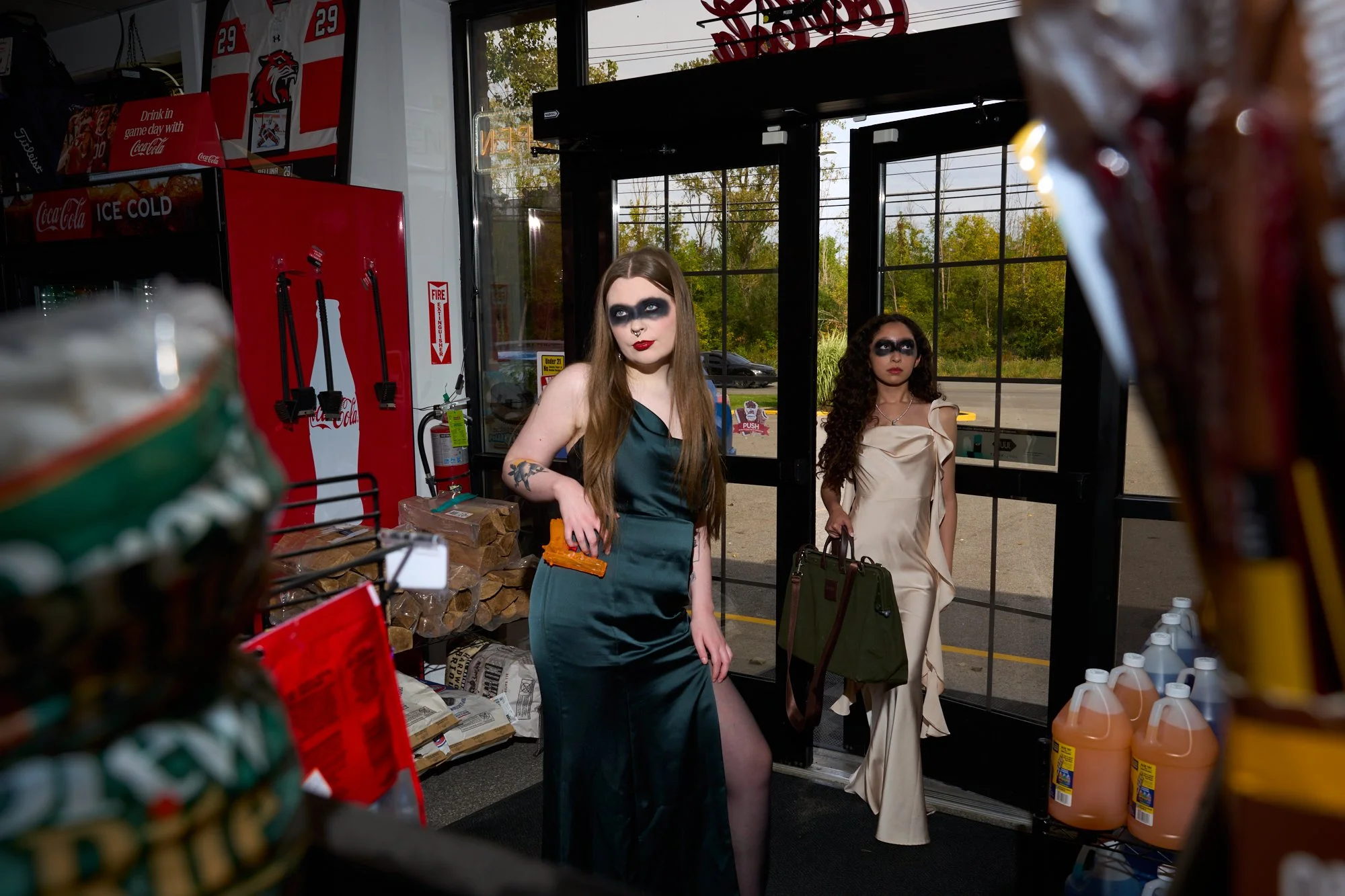 Two women with dramatic eye makeup and tattoos, one in a green satin dress and the other in a beige dress, entering a store through a glass door.  Snacks surround the two. One girl holds a green bag, and the other holds a bright orange water gun.