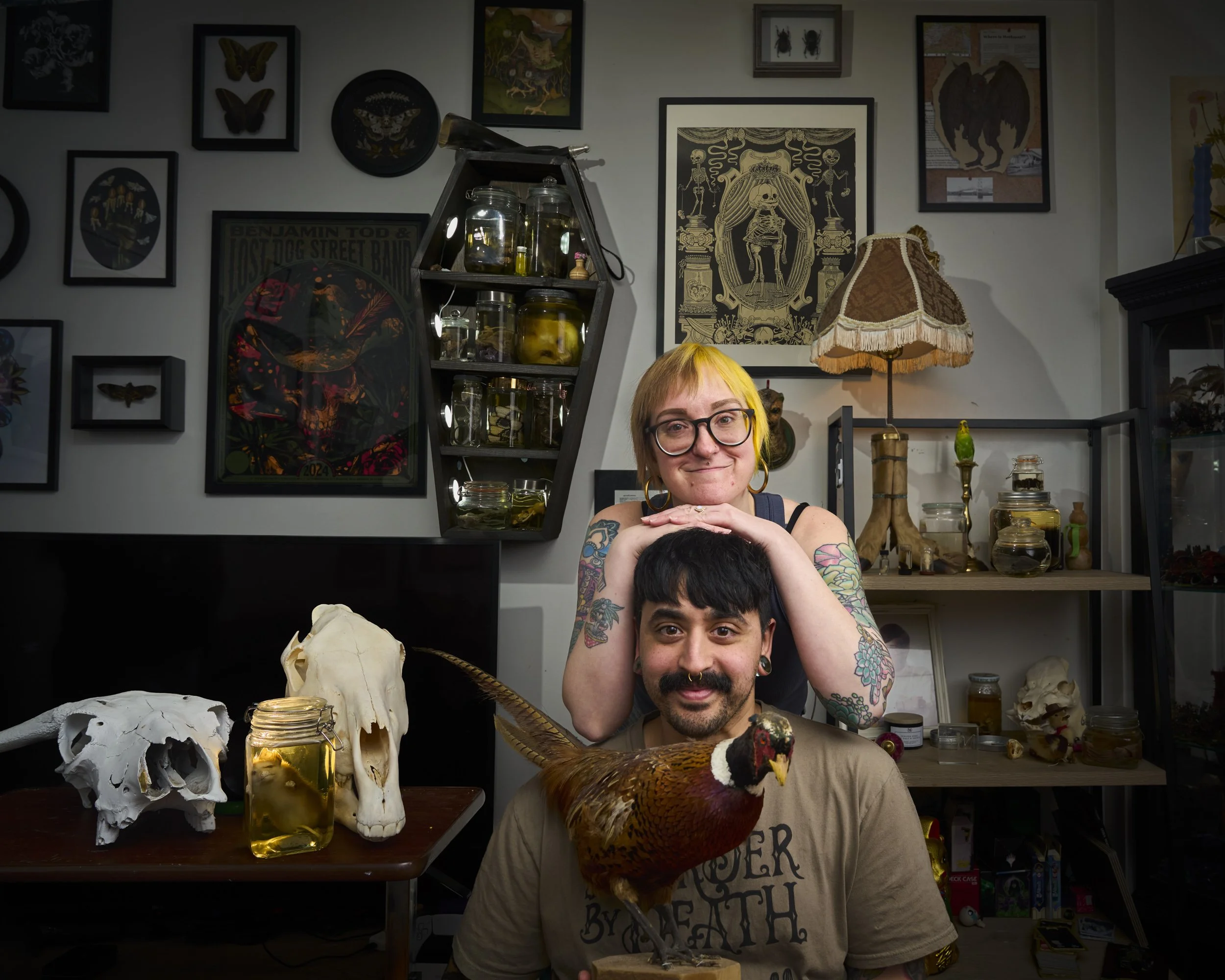 Two people, a woman with glasses and tattoos on her arms, and a man with a beard, are posing with a colorful pheasant bird on his shoulder in a room decorated with artworks, framed pictures, and various jars and animal skulls. The woman is smiling wi