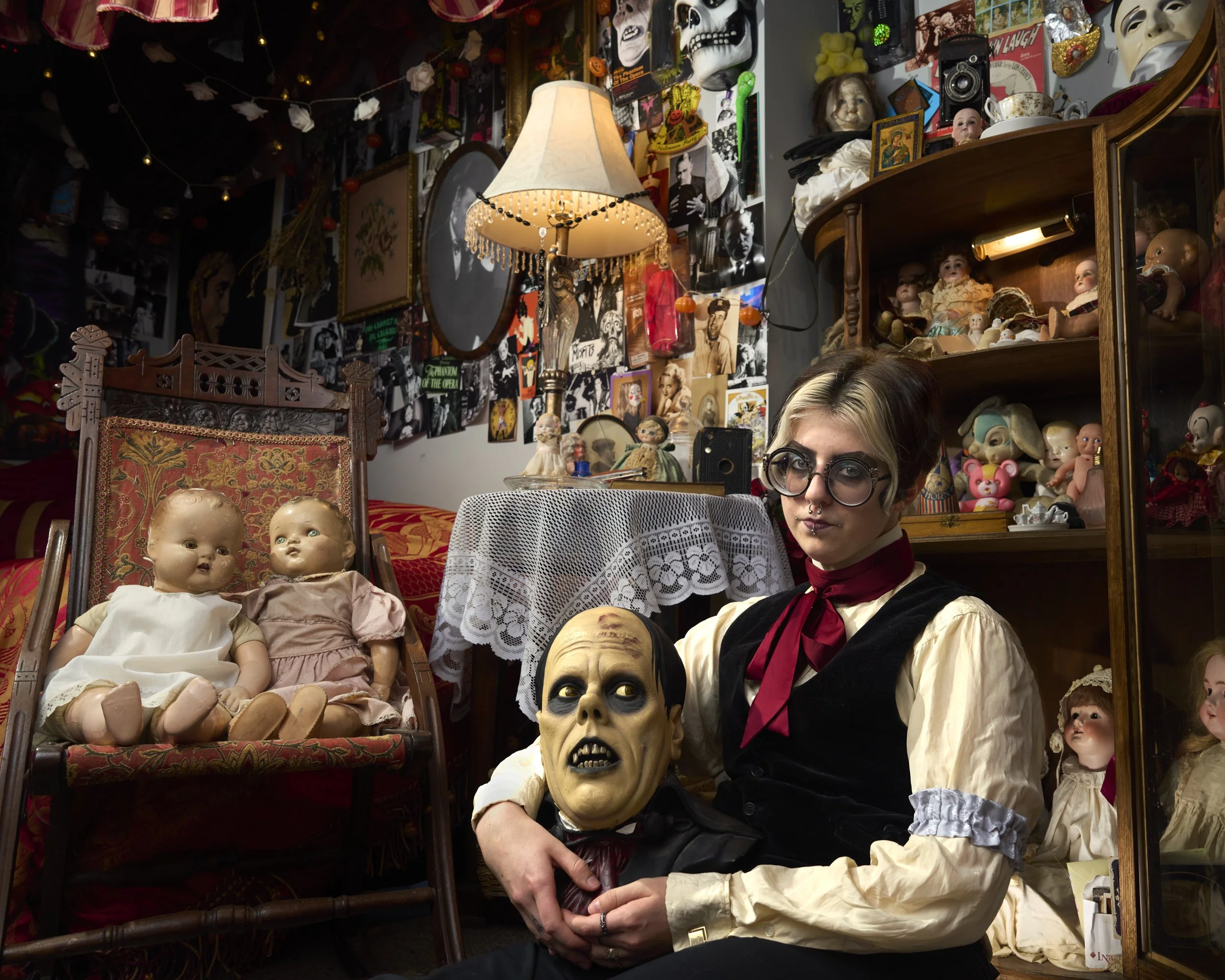 A person with short hair, glasses, and a serious expression sitting among vintage dolls and creepy masks in a room decorated with horror-themed posters and memorabilia.