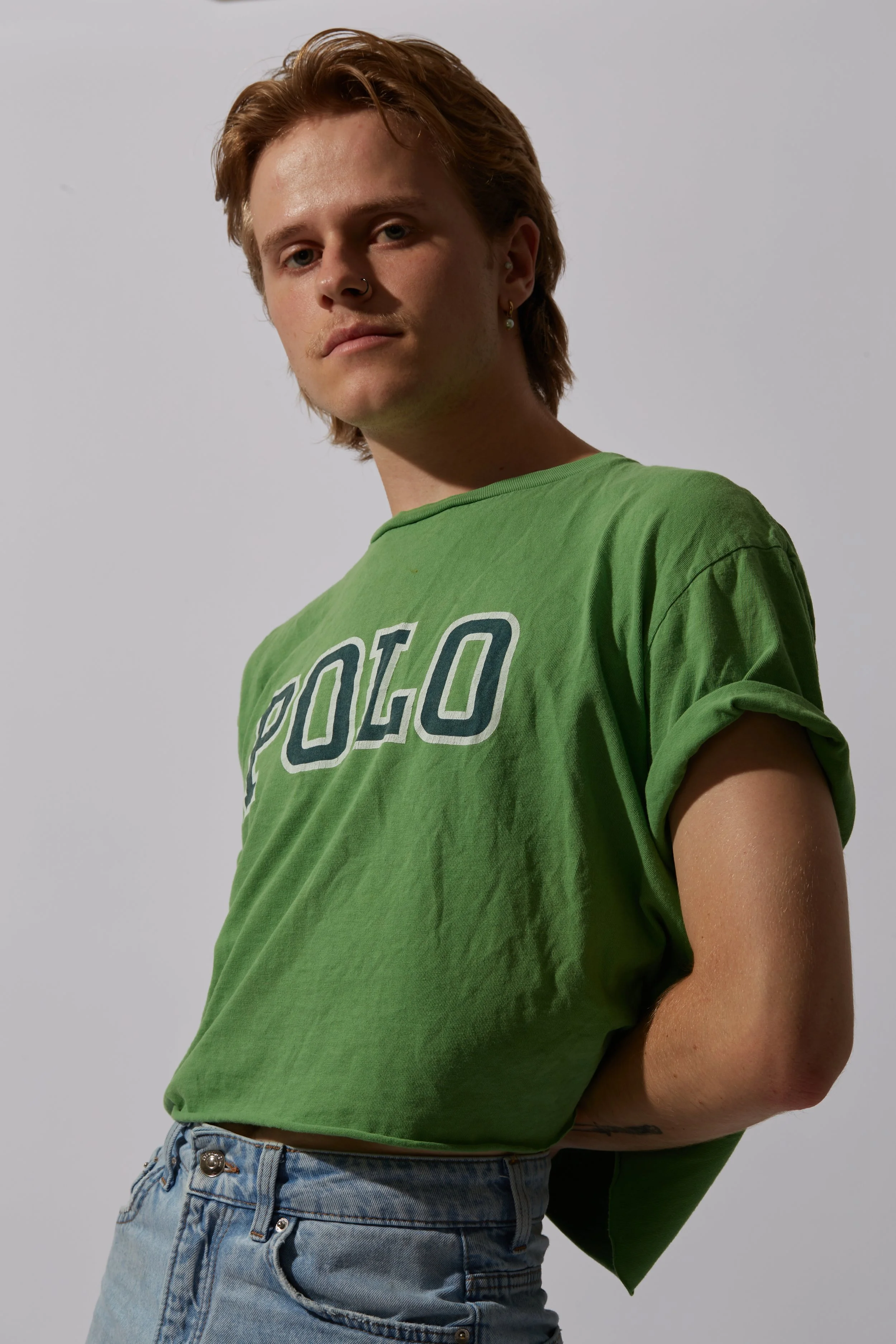 A young person with blond hair, multiple earrings, and a nose ring wearing a green t-shirt and denim jeans, standing against a plain white background.