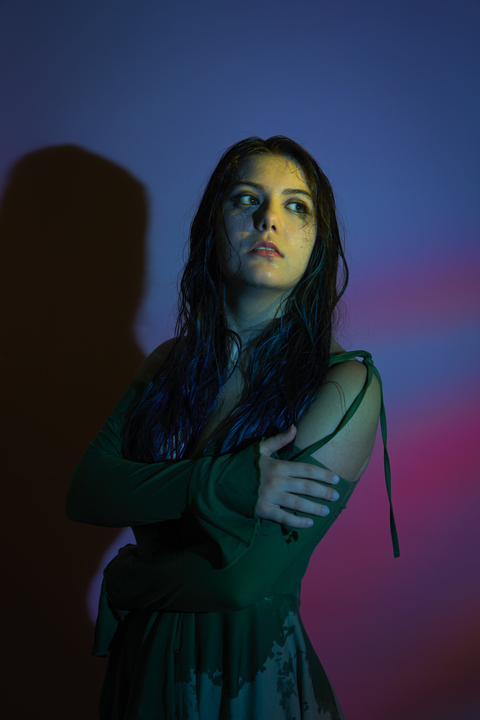 A woman with wet hair, wearing a dark green long sleeve dress, stands against a gradient purple and blue background. She looks to the side with a contemplative expression.
