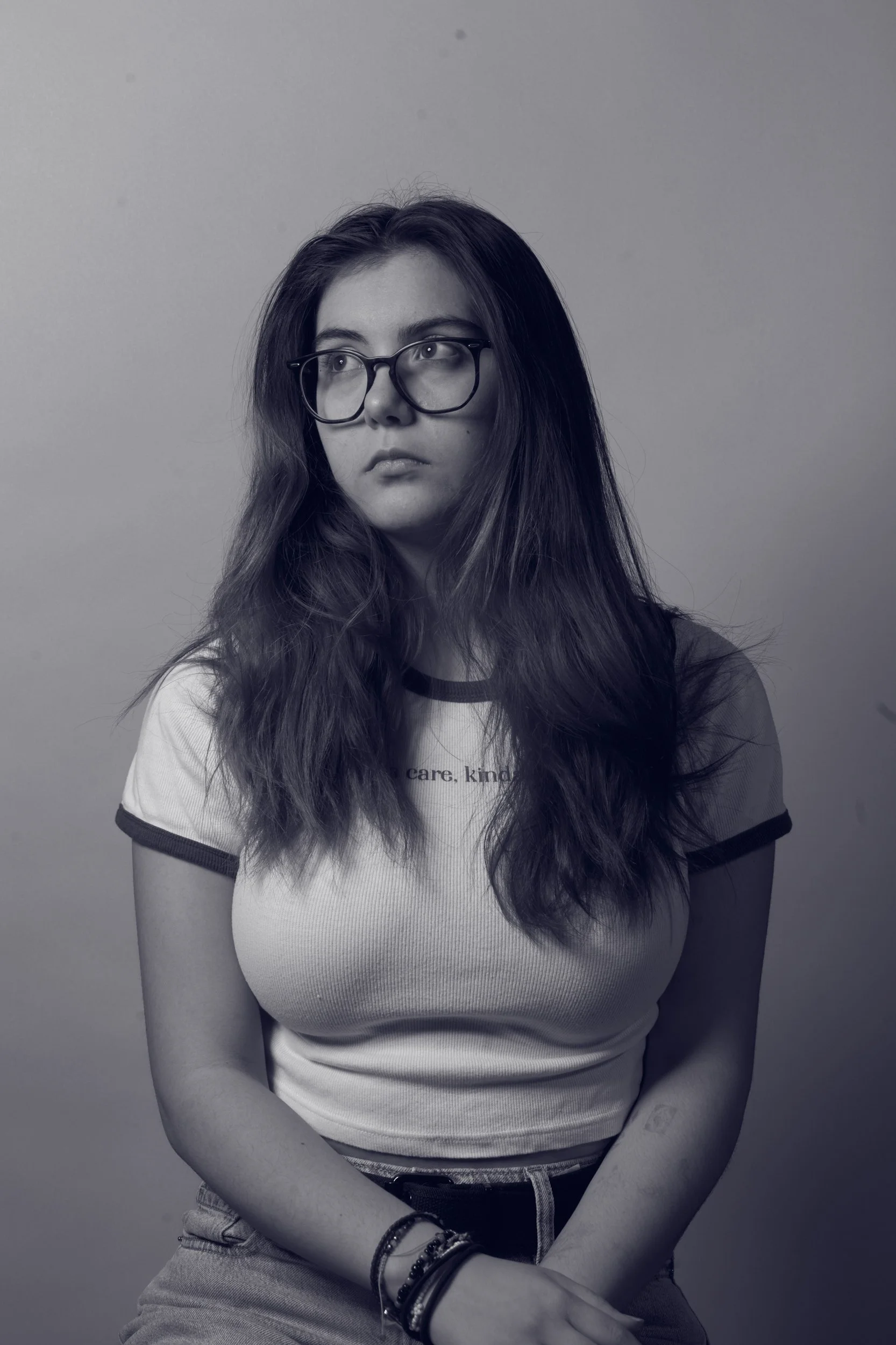Black-and-white photo of a young woman with long wavy hair, wearing glasses, a white t-shirt with dark trim, and light-wash jeans. She is looking off to her left with a neutral expression, seated with her arms crossed, against a plain background.