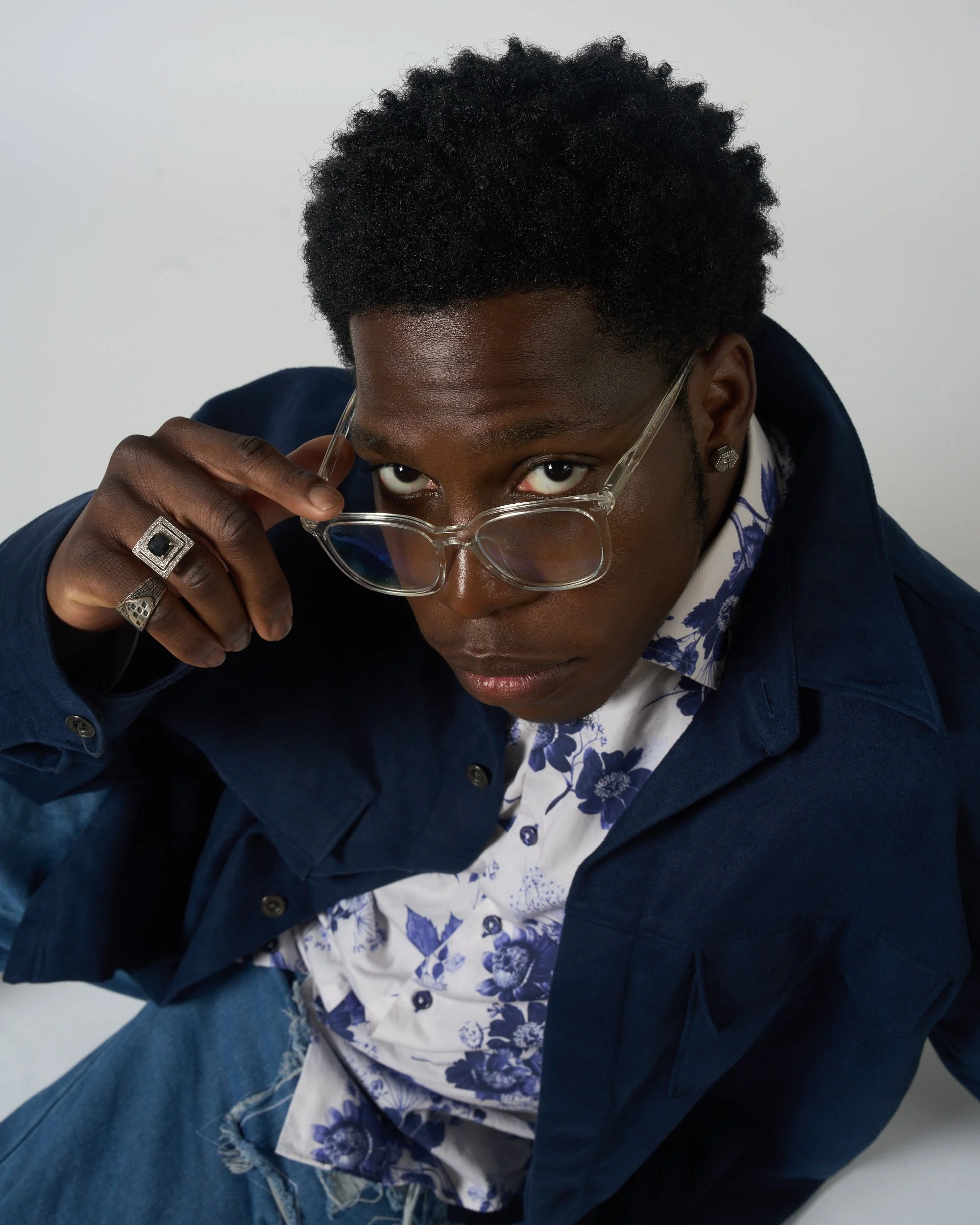 A young man with dark curly hair and dark skin looking over his glasses. He's wearing a dark jacket, a white shirt with blue flower patterns, and blue jeans. He has silver jewelry on his fingers and ears, and is adjusting his glasses with his right h