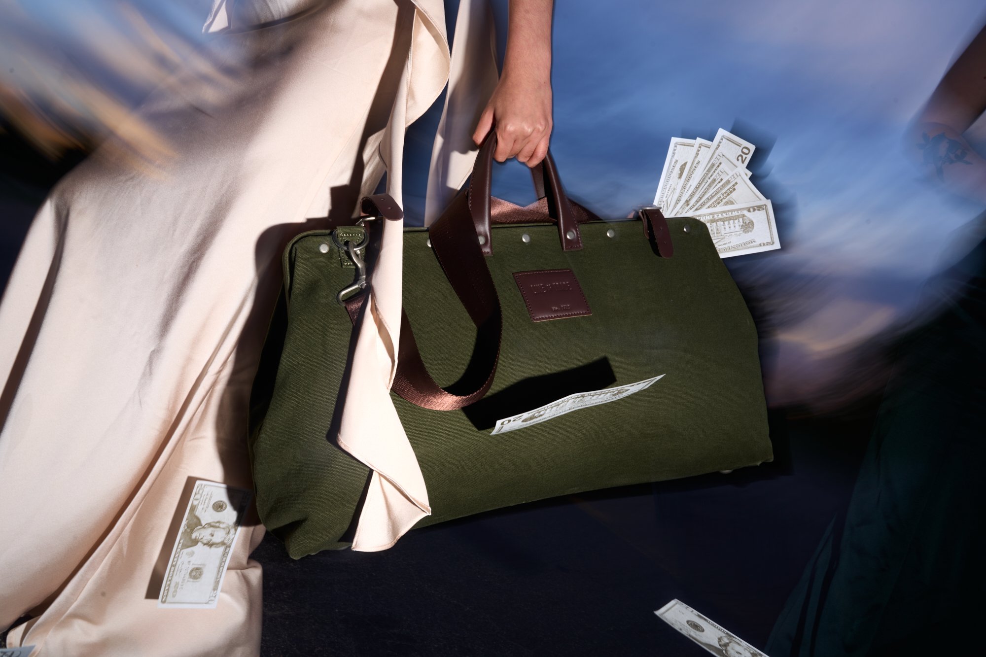 Close-up image of a person's hand holding a green handbag with brown handles, with US dollar bills partially visible inside and around it, on a dark surface.