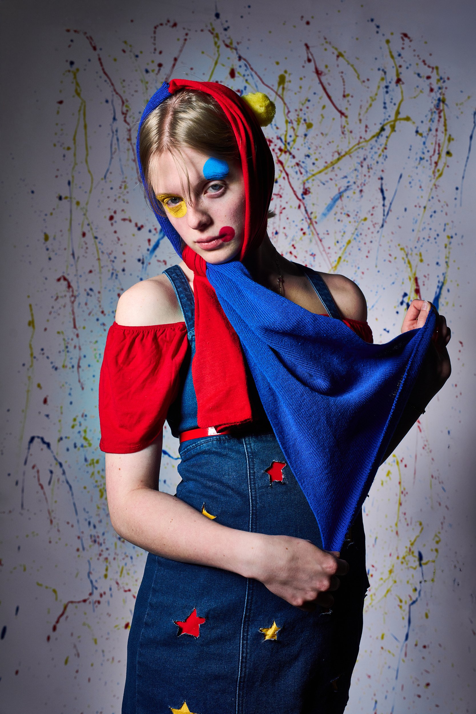 A young woman with painted circles on her face, wearing a red off-the-shoulder top, denim dress with star patches, and a hood made of red, yellow, and blue fabric. She is in front of a background with colorful paint splatters.