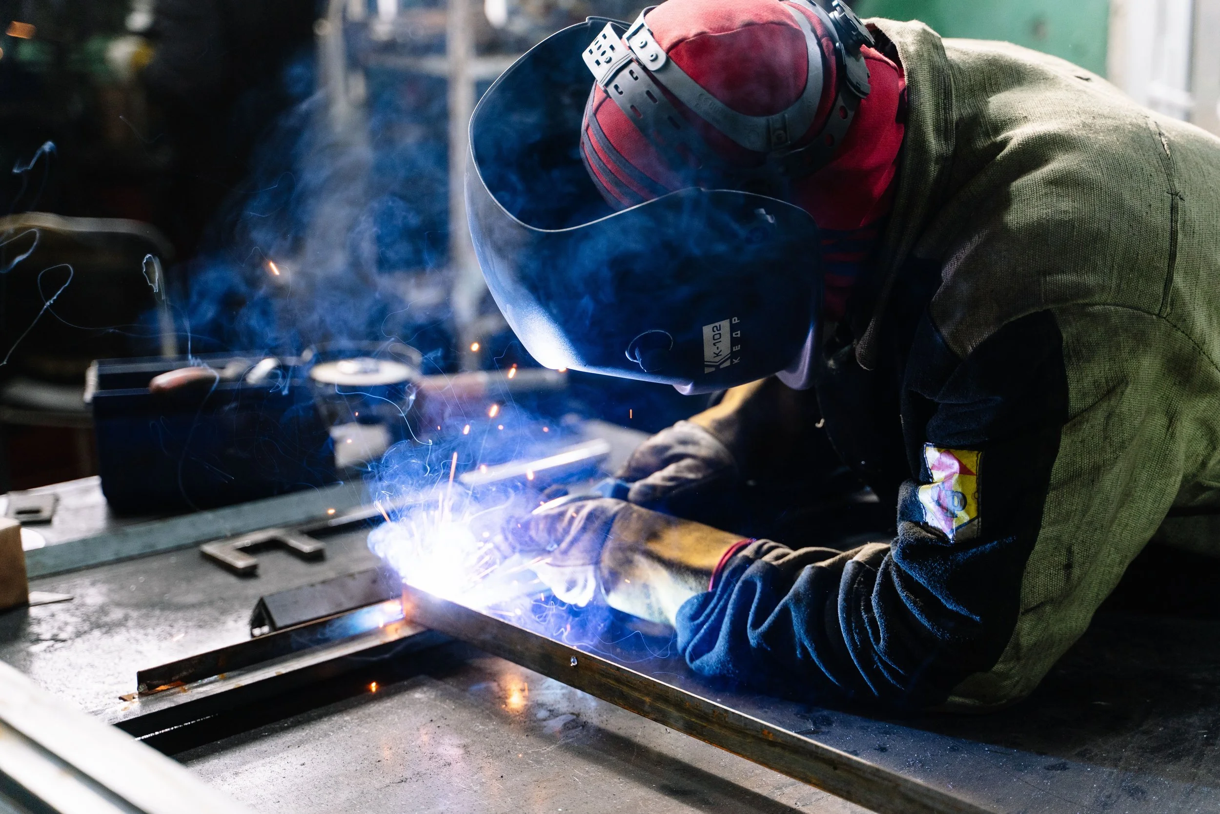 Welder wearing a protective helmet and gloves welding metal, with bright sparks and blue smoke.