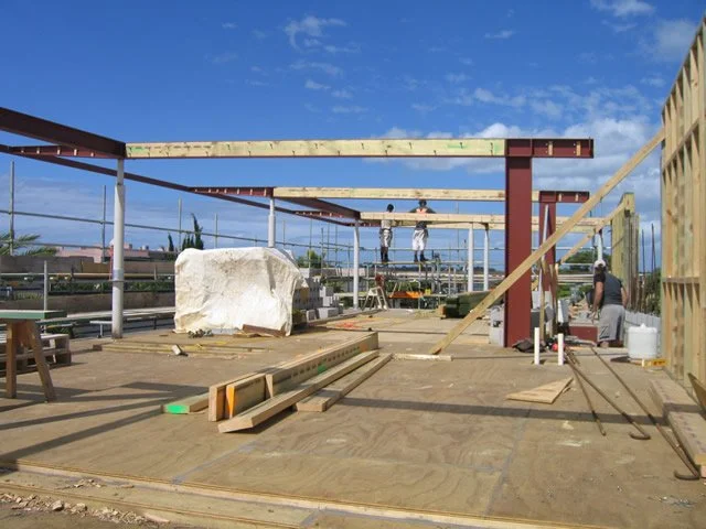 Steel-framed house under construction with workers, lumber, and building materials at an outdoor construction site