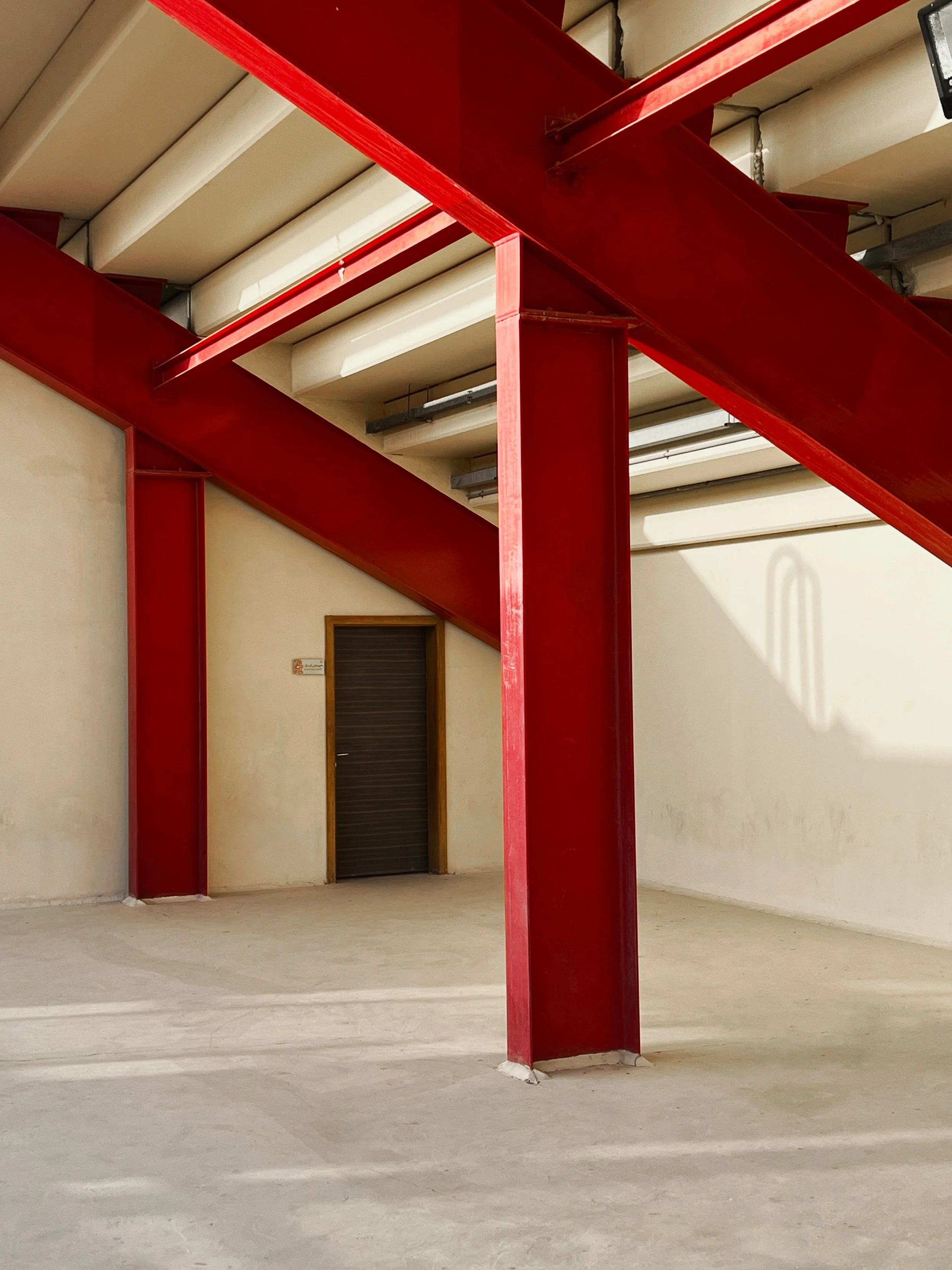 Interior of an industrial building with bright red steel support beams, white ceiling panels, concrete floor, and a dark door.