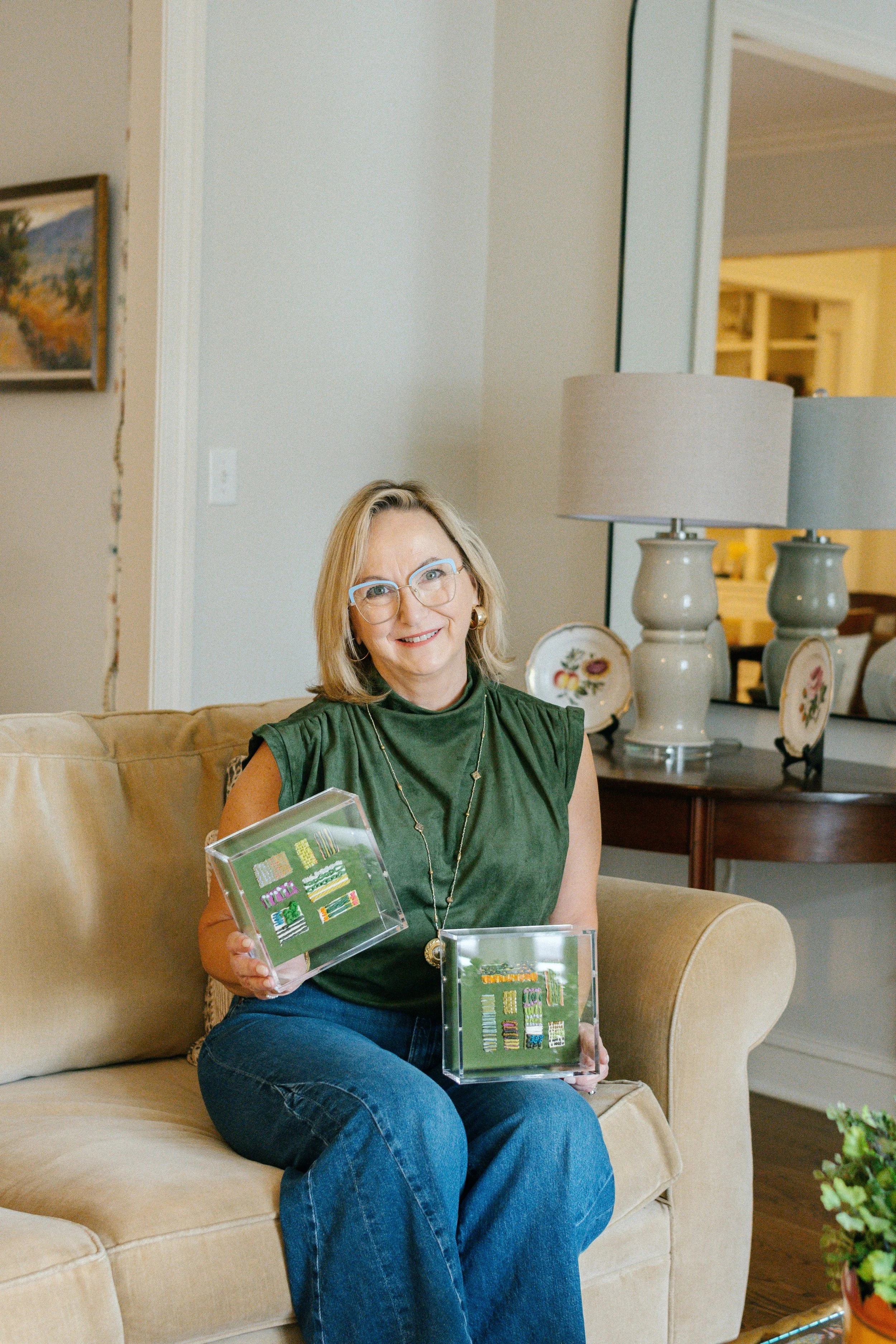 A woman with glasses and blonde hair sitting on a beige couch, holding a display case with various embroidered patches, smiling in a cozy living room with lamps, artwork, and decorative plates.