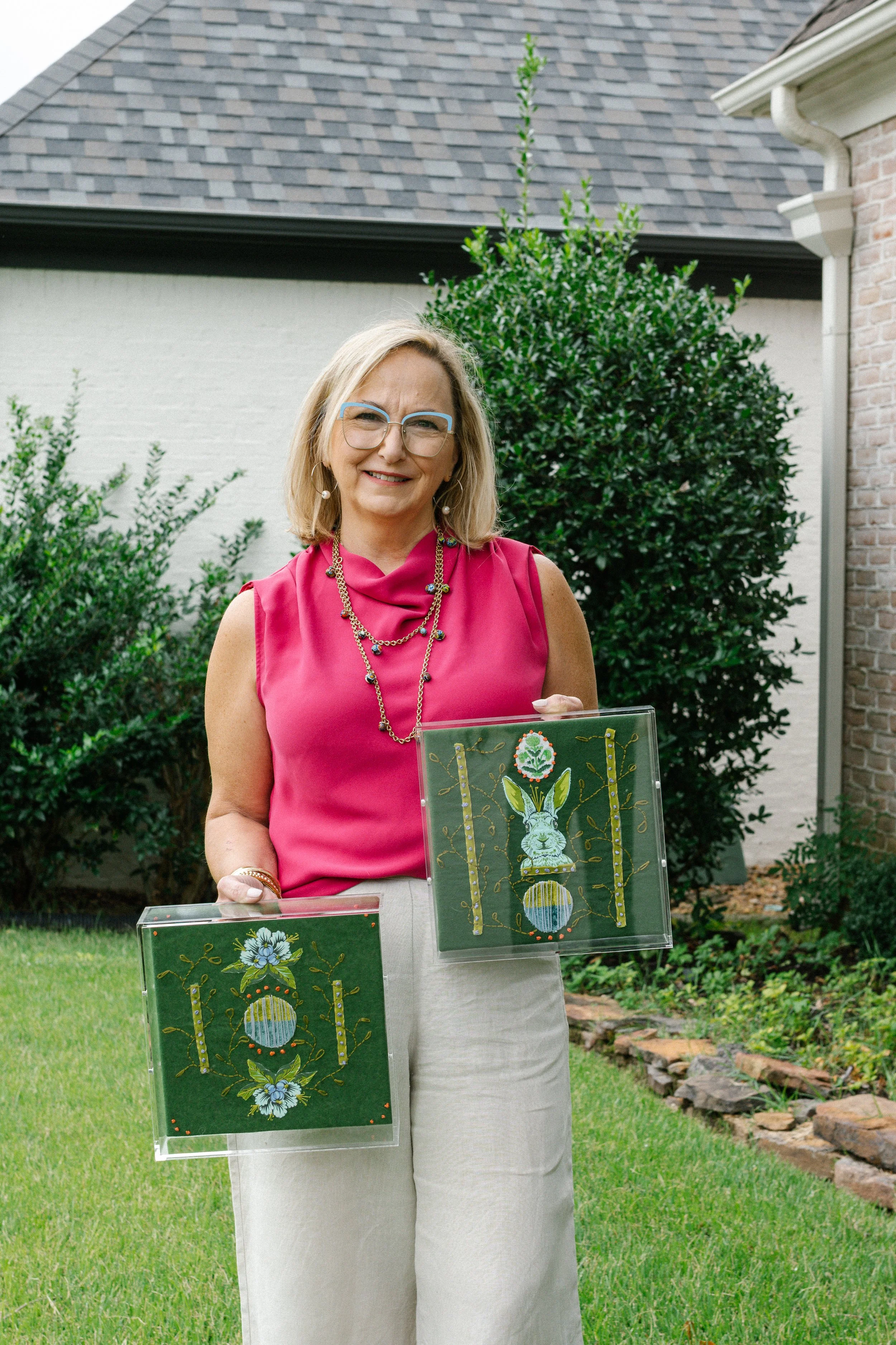 A woman with blonde hair, glasses, and wearing a pink sleeveless top and beige pants, standing outdoors in a garden holding two framed embroidered textile artworks. The background features green bushes, a brick house, a gray shingled roof, and a stone pathway.