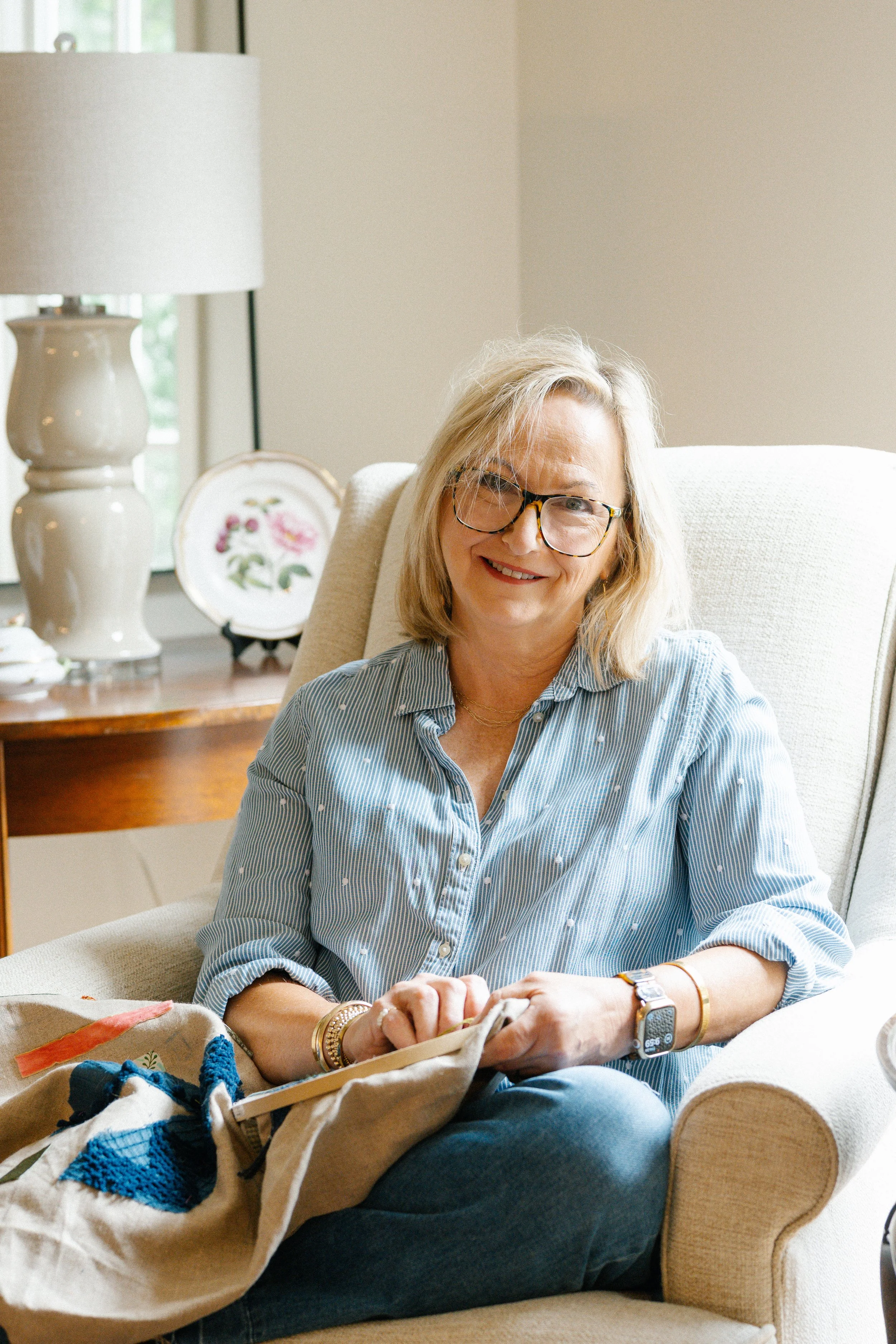 A woman sitting in a beige armchair, smiling, wearing glasses and a striped blue shirt, with knitting supplies on her lap. Behind her, a side table with paintings and a lamp is visible.