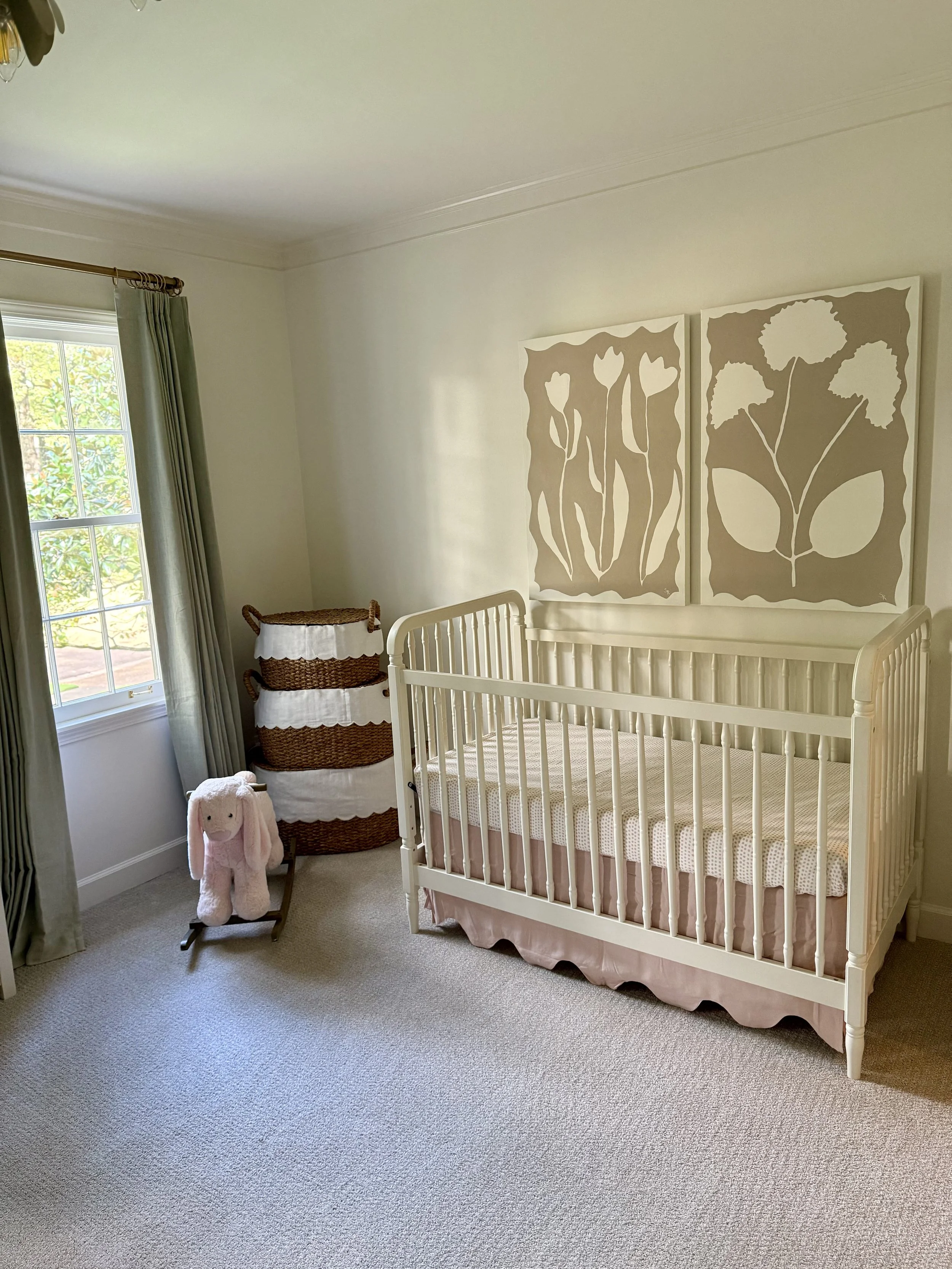 A nursery room with a white crib, two large beige and white botanical wall art pieces, a wicker laundry basket with a white cloth liner, a pink stuffed bunny toy, and a window with green curtains.
