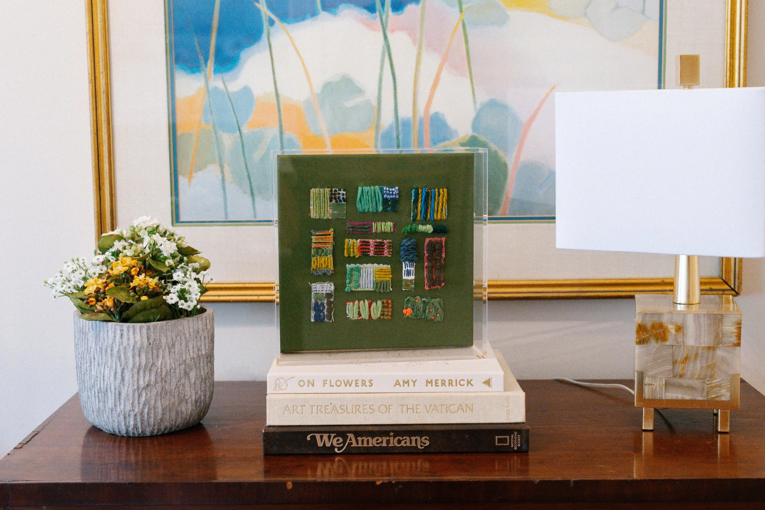 A wooden table with a potted plant with white and yellow flowers, a set of books stacked with titles including "On Flowers" and "Art Treasures of the Vatican," a green display case with colorful yarn samples, a white table lamp with a gold base, a small gold rectangular object, and a large abstract watercolor painting with blue, orange, green, and pink hues in a gold frame in the background.
