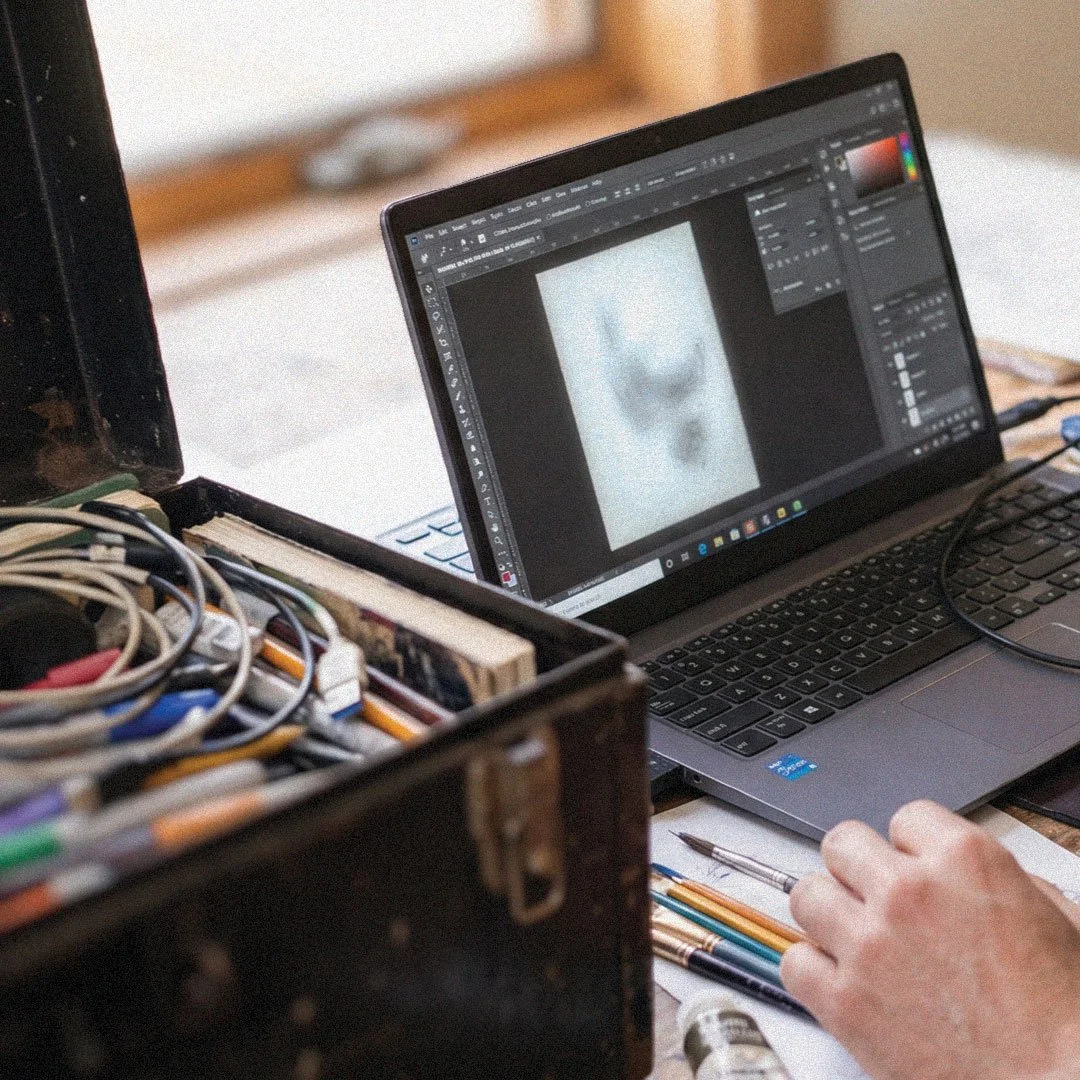 Artist editing a digital artwork on a laptop beside a box of paintbrushes and art supplies