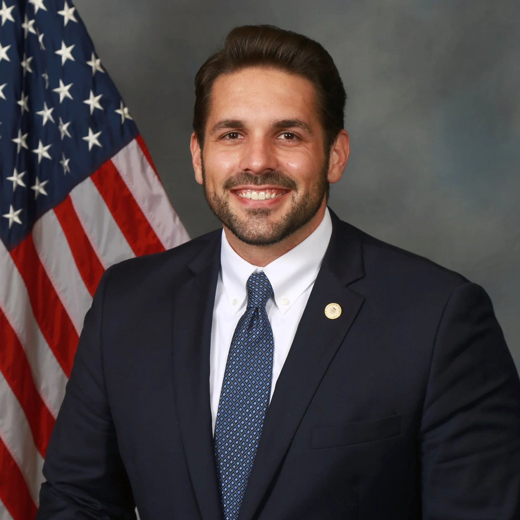 Mayor Scott Conger in a dark suit, white shirt, and blue patterned tie, sitting in front of an American flag, smiling at the camera.