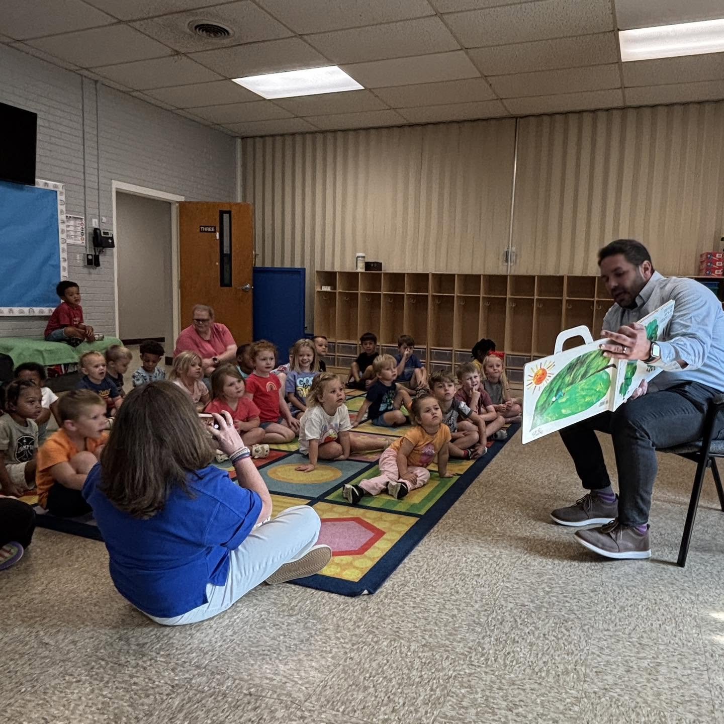 Mayor Scott Conger reading a colorful children's book to a group of young children sitting on a carpet in a classroom