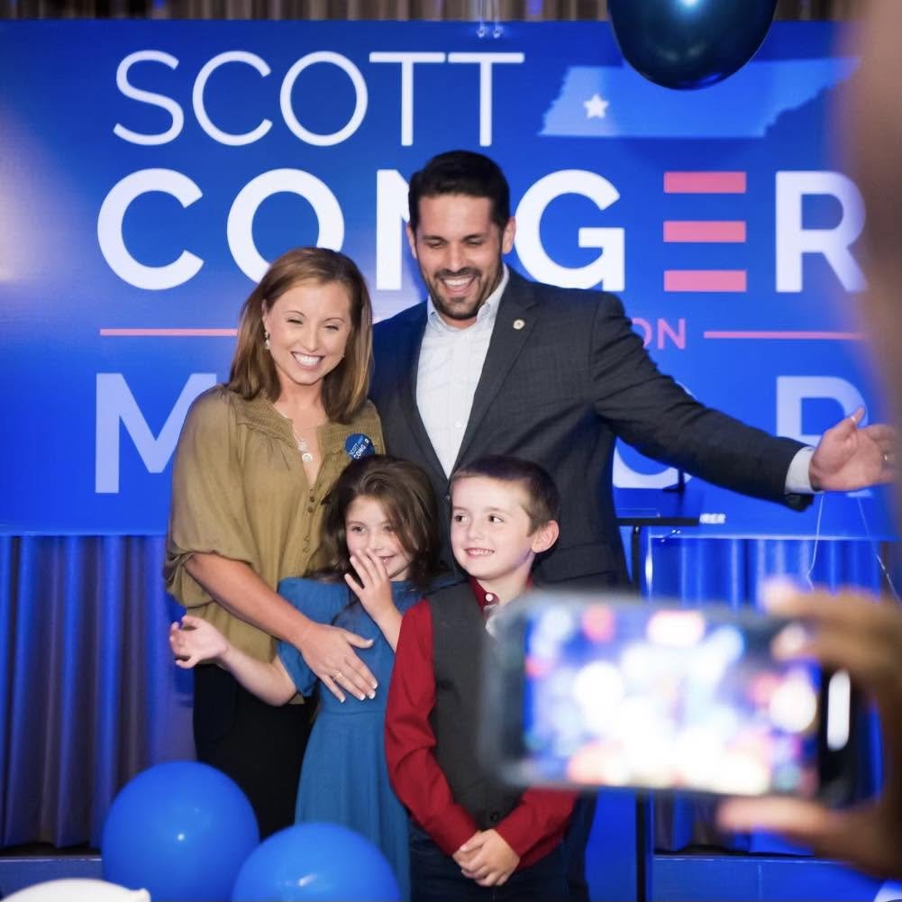 Scott Conger celebrating with his wife and two children at his previous campaign event with blue balloons and a campaign sign in the background.