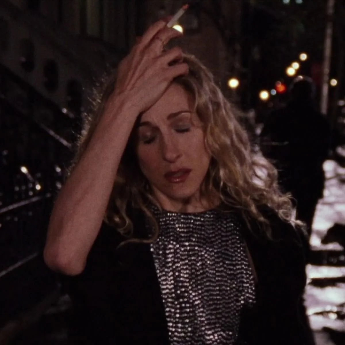 A woman with curly blonde hair, eyes closed, touching her forehead in a dimly lit outdoor setting at night, with city lights in the background.