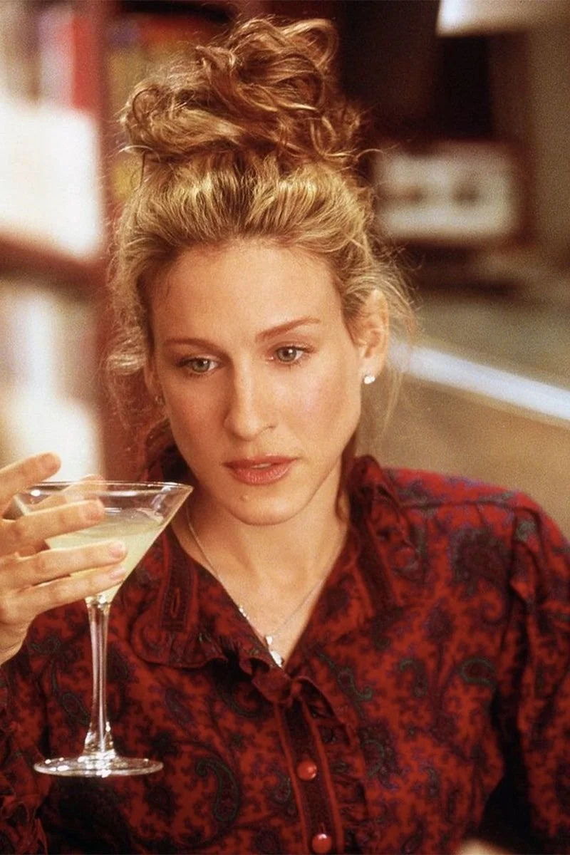 A woman with curly red hair styled in an updo, wearing a red patterned blouse, holding a cocktail glass, and looking at it thoughtfully.