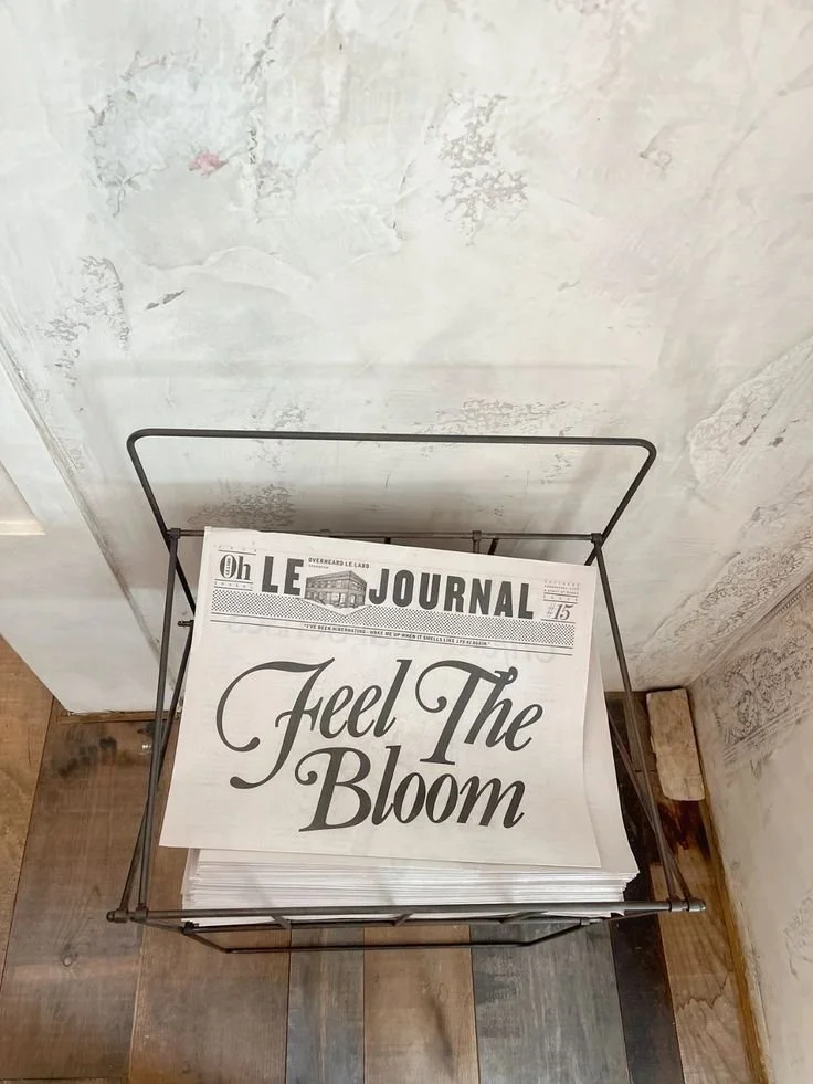 A stack of newspapers in a black wire basket. The visible front page has the title 'The LE JOURNAL' and the headline 'Feel The Bloom' in large cursive letters. The background consists of a textured, partially painted wall and a wooden floor.