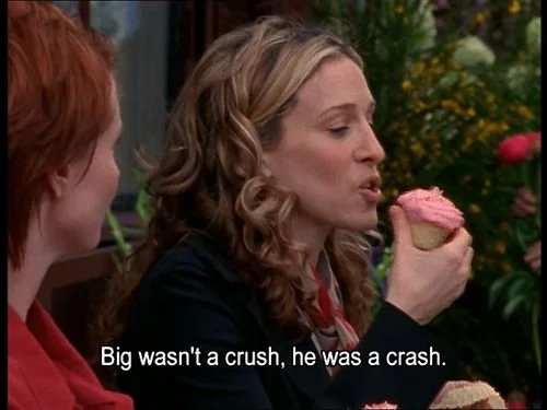 Woman with curly blonde hair holding a cupcake with pink frosting, speaking to another woman with red hair.