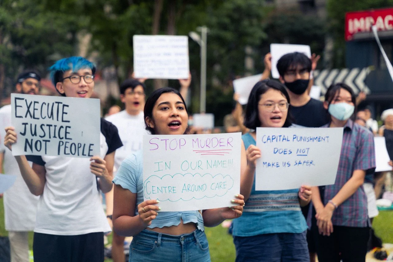 Group of diverse protesters holding signs at a rally, advocating for justice, ending murder, and opposing capital punishment, outdoors in a park.