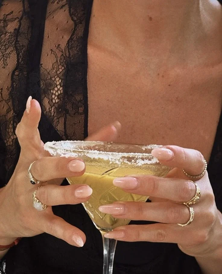 Close-up of a woman holding a margarita glass with a salted rim, wearing multiple rings and a black lace garment.