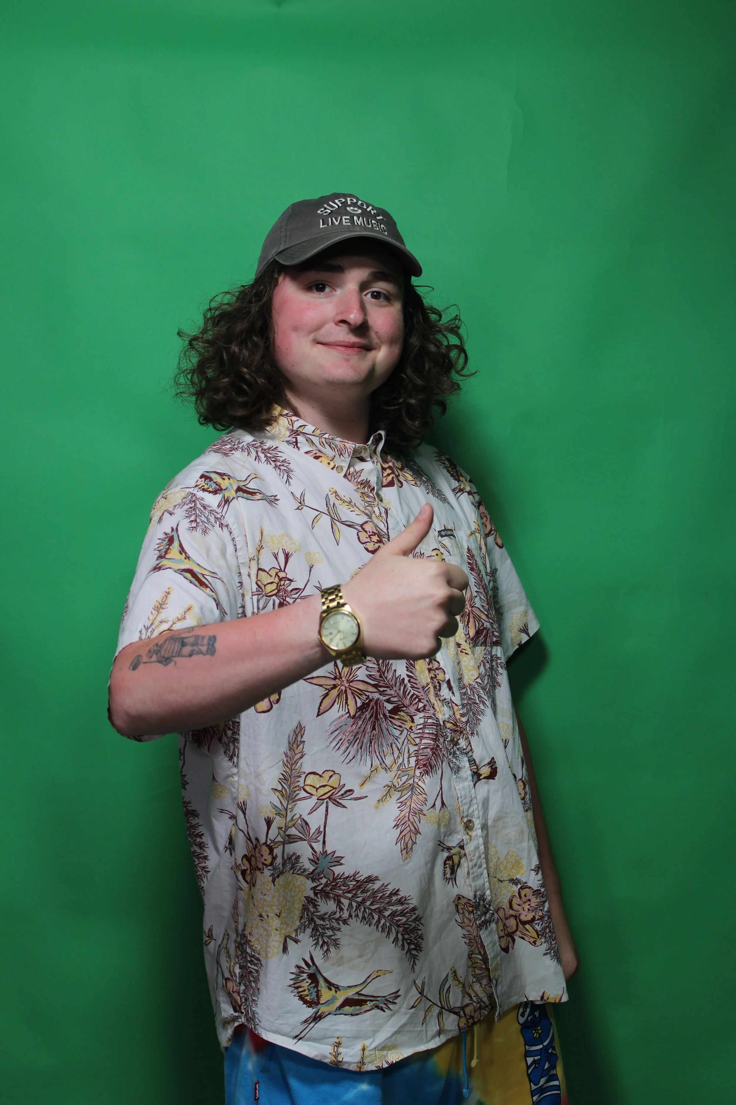 A young man with curly hair and a tattoo on his left arm, wearing a floral-patterned shirt, a gold watch, and a black hat with the words "Support Live Music" written on it, giving a thumbs-up gesture against a green background.