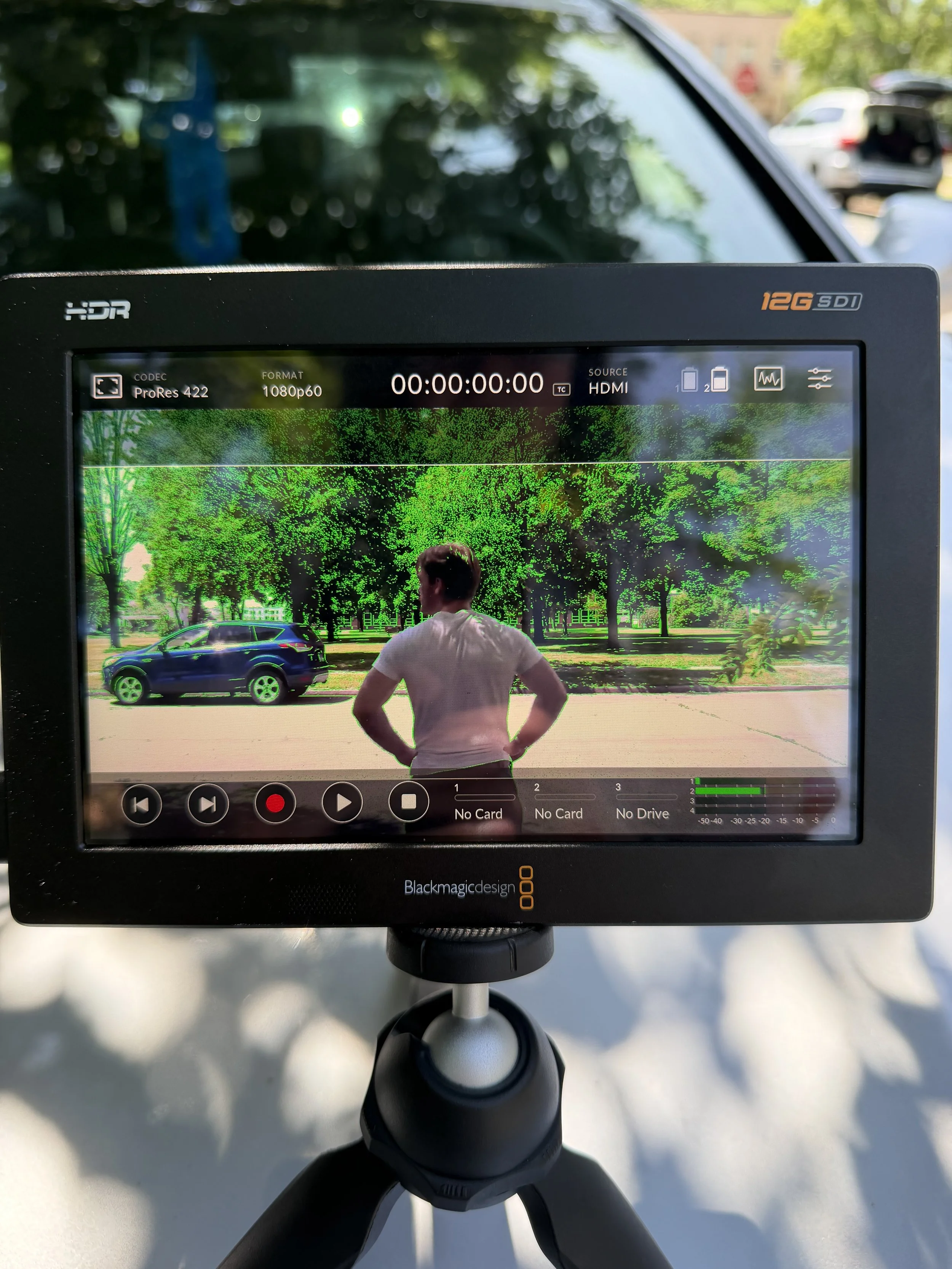 A professional monitor displaying a recording of a man in a white t-shirt with hands on hips, standing outdoors near a black car, with green trees in the background.