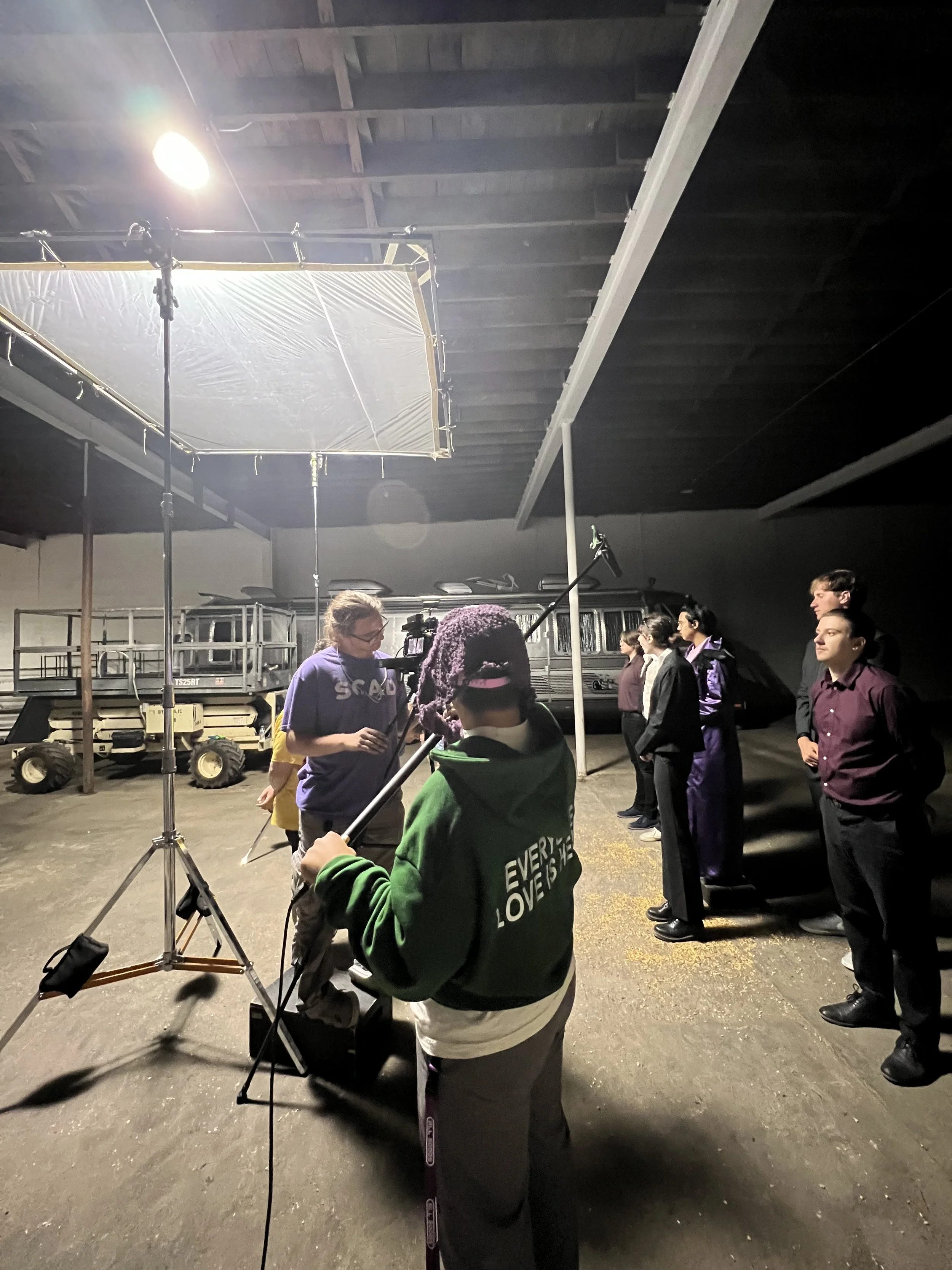 A group of people standing in line inside a warehouse-like space, preparing for a production or filming. There is lighting equipment and a tripod with a camera set up, with a crew member adjusting it. The people are wearing casual and business casual attire, and the setting appears to be a behind-the-scenes shot of a media production.