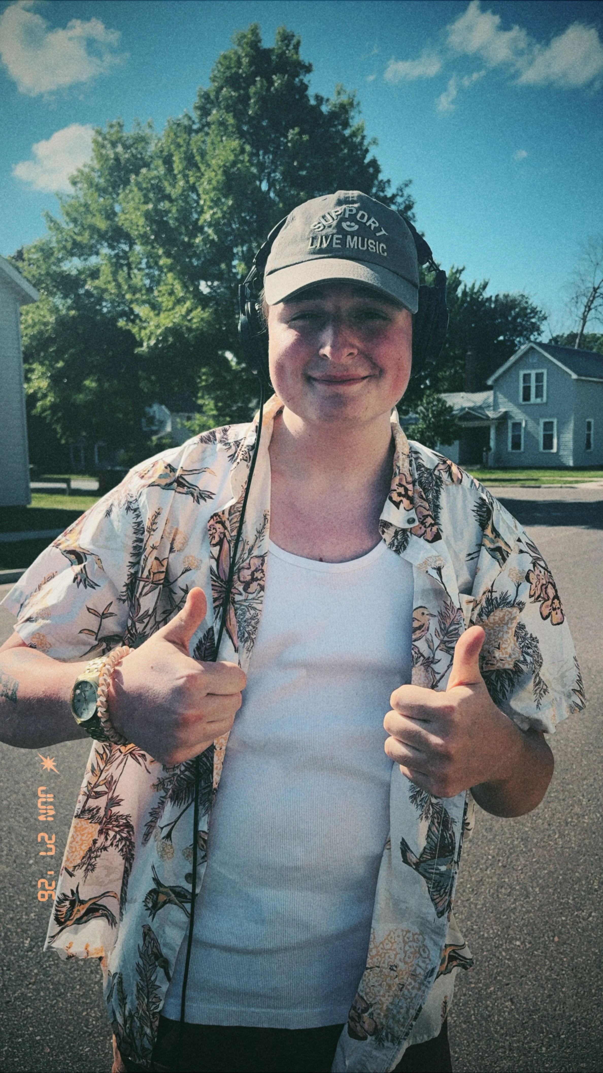 A person wearing a gray cap with 'Support Live Music' embroidered on it, listening to headphones, smiling with a thumbs-up gesture, wearing a white tank top and a floral-patterned short-sleeve shirt, standing outdoors on a sunny day with trees and houses in the background.