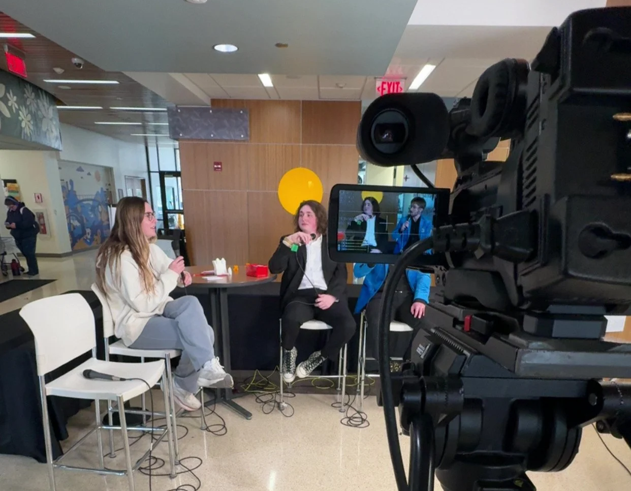 Two women being recorded during an interview in a public indoor space, with a professional camera in the foreground.