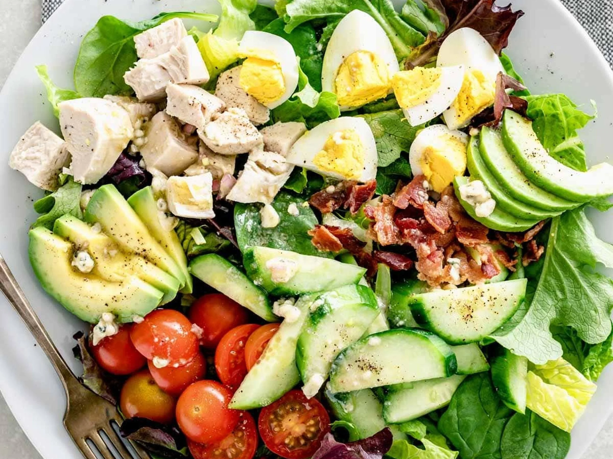 Mixed salad with cherry tomatoes, cucumber, avocado, boiled eggs, grilled chicken, bacon bits, leafy greens, and blue cheese crumbles.